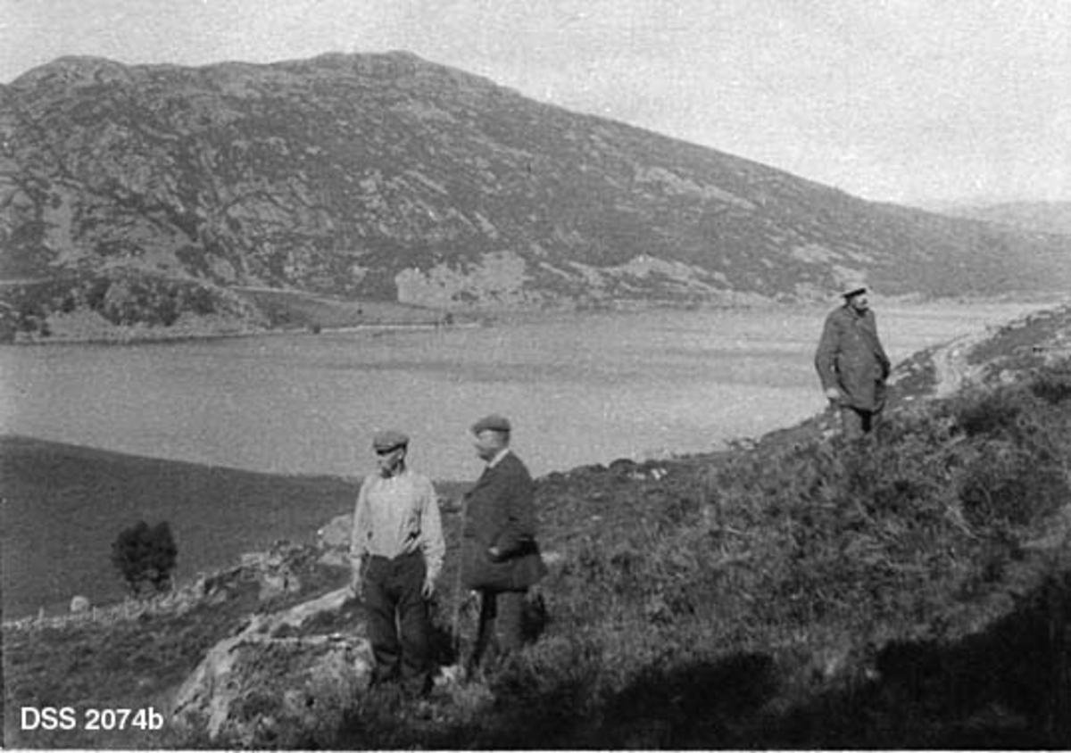 Utsikt fra Røyslandfjell på Jæren.  Todelt landskapsopptak, tatt mot nedenforliggende flate med vatn og jordbruksareal.  En veg later til å krysse vatnet over et smalt eid som later til å være en sandbanke.  Gardene på motsatt side av vatnet heter Røysland.  Bakenfor gardene igjen er det en kraftig bergrygg med lite vegetasjon.  Bildet er sammensatt av to opptak.  I forgrunnen på det høyre opptaket står tre menn på lyngmarka og skuer ut over landskapet. 