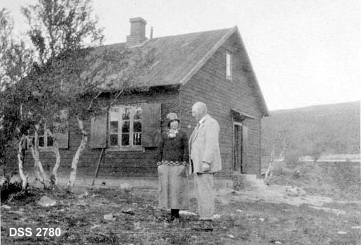 Avsjøhytta på Dovrefjell, bordkledd bindingsverkshus i fjellandskap med bjørketrær.  Mann og kvinne foran hytta, muligens ekteparet Ielstrup. 