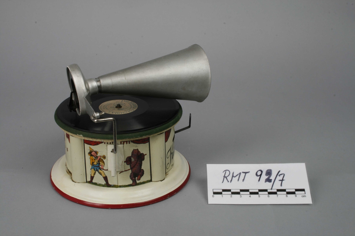 Leketøysgrammofon for avspilling av plater. Fjærmekanikk innvendig som trekkes opp for avspilling.
Nål og trakt som høytaler. 
Alle deler er i metall. Topplaten er malt grønn og limt rett på metall. Muligens en slags vinylmaling.