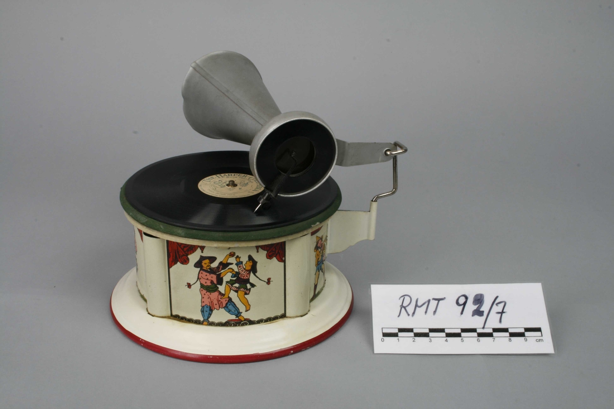 Leketøysgrammofon for avspilling av plater. Fjærmekanikk innvendig som trekkes opp for avspilling.
Nål og trakt som høytaler. 
Alle deler er i metall. Topplaten er malt grønn og limt rett på metall. Muligens en slags vinylmaling.