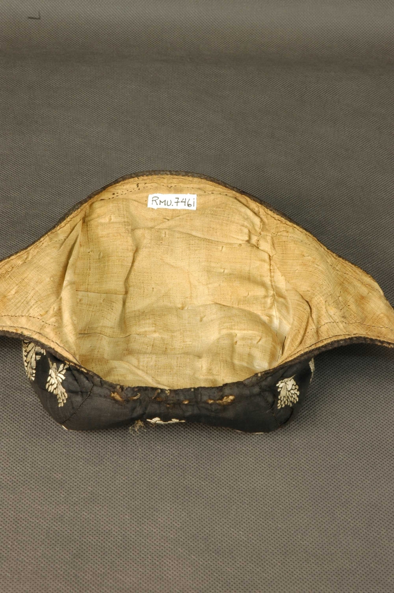 Ørelue i svart silke med hvite broderte roser. Ingen dusk. Foret med lyst linstoff. Svarte knytteband.
