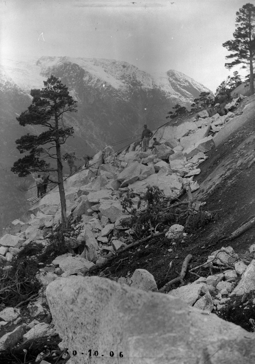 Anleggsarbeidere henger i tau på enkle stillas i fjellsida.