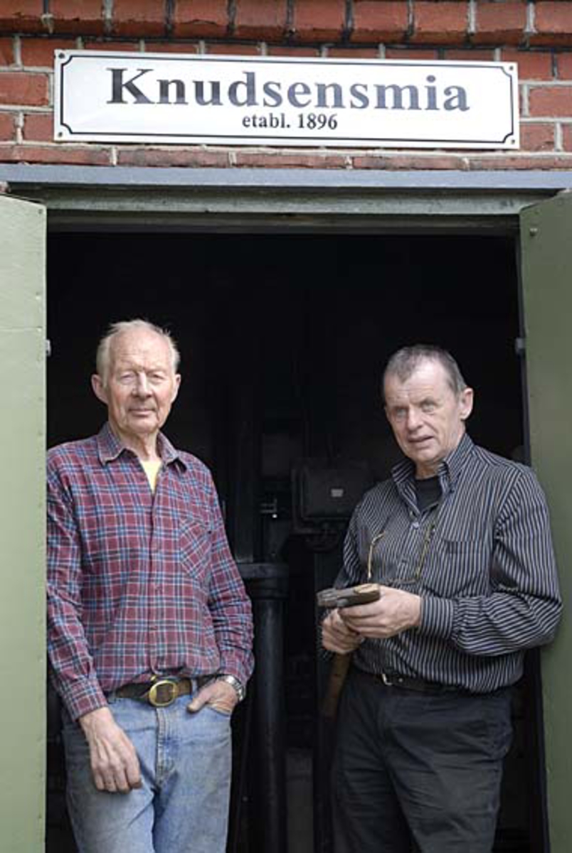 Fra Knudsen-smia i Mjøndalen.  Fotografiet er tatt ved inngangsdøra med [Karl] Oddbjørn Knudsen (f. 1935) til venstre og John Lystad (f. 1945) til høyre.  Oddbjørn er tredje generasjon smed i familiebedriften, som produserte merkeøkser, strafføkser (avkortingsøkser), blinkeøkser, hoggøkser og fløterhaker i Mjøndalen i perioden fra 1896 til 1980.  Oddbjørn begynte i lære hos faren Odmund (f. 1900) i slutten av 1940-åra, og hadde fulltidsjobb i smia fram til 1963-64.  Deretter hjalp han faren, som holdt virksomheten gående til 1980, når han hadde ledig tid.  Oddbjørn har restaurert smia, og som pensjonist driver han formidling av håndverkstradisjoner sammen med assistenten John Lystad.  Skiltet over hodet på karene er produsert i København i forbindelse med at smia ble restaurert. 