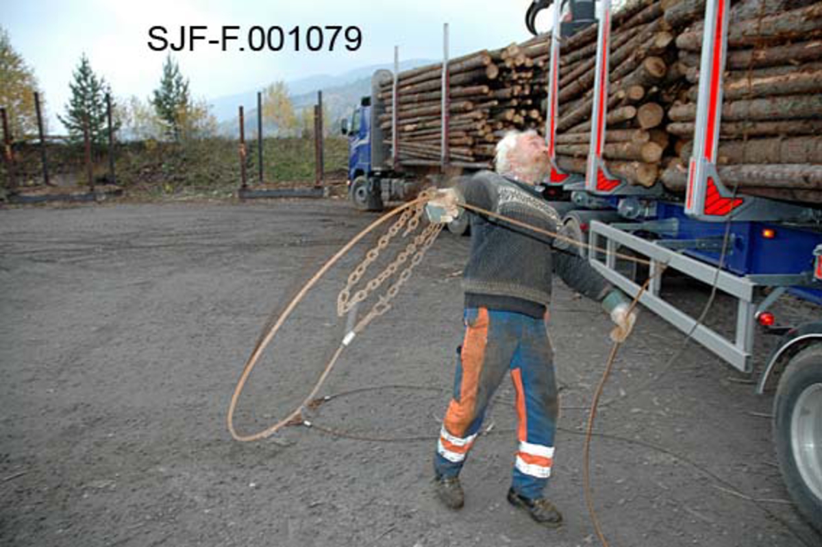 Fra Skiensvassdragets fellesfløtningsforenings utislagsplass på Nesøya ved Notodden.  Fotografiet viser Tom Kaafjeld, en av medarbeiderne i Skiensvassdragets fellesfløtningsforening, idet han kaster et "bilbind" over et lastebillass med tømmer. Et bilbind besto av cirka ti meter vaier og cirka to meter med kjetting.  I enden av kjettingen var det et pæreformet lås.  Som en sikkerhetsforanstaltning ble låsene surret med tynn stålståd, såkalt blomstertråd.  Bilbindet skulle følge tømmeret fra utislaget til det var fløtet fram til papirfabrikken.  

Kaafjeld er iført svart strikkegenser med kvitt mønster og oransje og blå arbeidsbukser.  Han beskytter hendene ved å bruke arbeidshansker. 