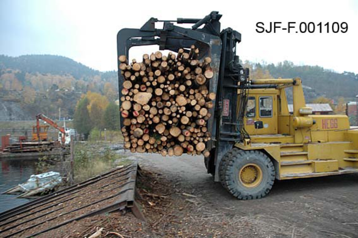 Fra Skiensvassdragets fellesfløtningsforenings utislagsplass på Nesøya ved Notodden.  Fotografiet viser utislag av buntet tømmerlass fra en 32 tonns gaffeltruck.  Den digre, gule kjøredonigen med et tømmerlass i "klørne" er på veg mot utislagsrampa, et skråplan som består av parallelle stålskinner, og som fører ned mot vannkanten.  Bunten skal slippes nedover på disse skinnene.  Deretter samles og bindes denne og andre bunter sammen til et større slep. 