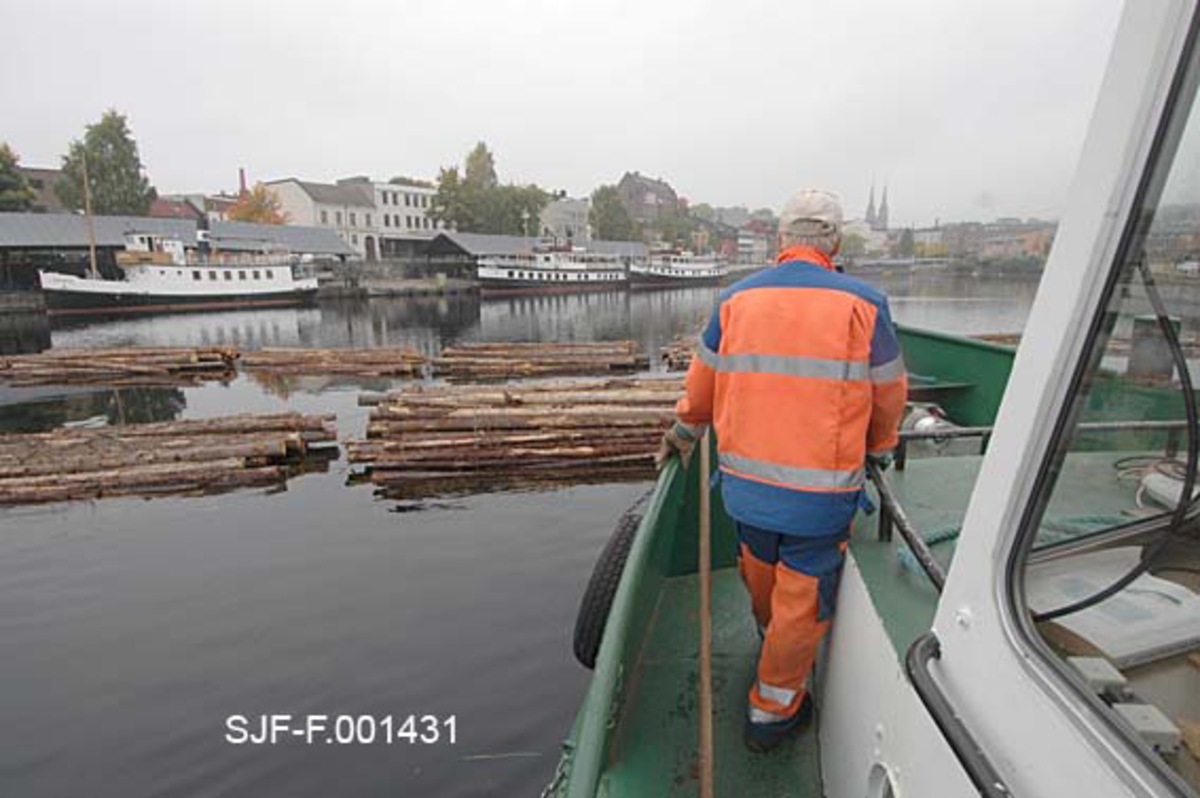 Torgrim Mork på dekket av slepebåten Triset.  Vi ser noe av styrhuset.  I bakgrunnen ligger et tømmerslep på vannet, og ved brygga på motastt side ligger tre veteanbåter.  Fotografiet skulle forakres ved land på Hjellevannet, ved den såkalte Damfossen, i påvente av at en kollega fra Skiensvassdragets fellesfløtingsforening med en mindre båt kom for å hente kortere lenker med tømmer for slusing gjennom Skien sluse til det nedenforliggende Bryggevannet og papirfabrikken Union. 