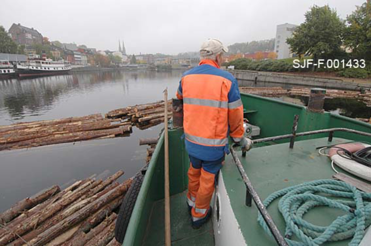 Torgrim Mork på fordekket av slepebåten Triset.  Mork står og forsøker å få overblikk over lenkene med tømmerbunter som ligger i vannet ved båten.  Ved siden av seg har han en fløterhake.  Mork forbereder seg på å klatre ned på tømmeret for å kunne binde det godt ihop.  Fotografiet er tatt på Hjellevannet, ved den såkalte Damfossen, der tømmeret ble lagt i påvente av at en kollega fra Skiensvassdragets fellesfløtingsforening med en mindre båt kom for å hente kortere lenker med tømmer for slusing gjennom Skien sluse til det nedenforliggende Bryggevannet og papirfabrikken Union. 