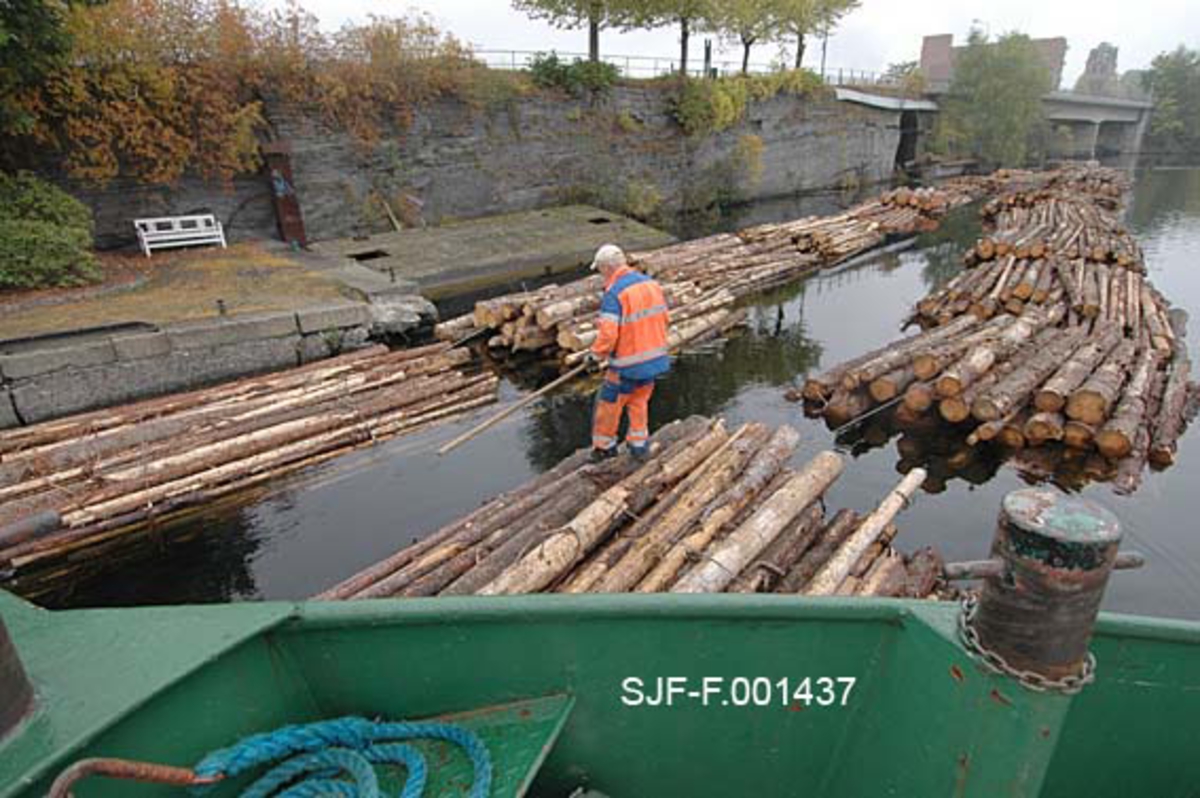 Torgrim Mork med fløterhake på tømmerbuntene på Hjellevannet, ved den såkalte Damfossen.  Her ble tømmeret, som var fløtet fra Notodden gjennom Løveid sluser og nedover, lagt mot land i påvente av at en kollega fra Skiensvassdragets fellesfløtingsforening skulle komme med en mindre båt kom for å hente det til slusing.  Slusetømmeret ble "satt inn" i Skien sluse i kortere lenker.  Derfra fløt de nedover Bryggevannet et stykke til de nådde tømmermottaket på papirfabrikken Union.  I fronten ses baugspissen på slepebåten Triset, og vi aner at båten ble brukt til å dytte lenkene med tømmerbunter mot hverandre slik at virket ble komprimert og ikke var til sjenanse for annen båttrafikk. 