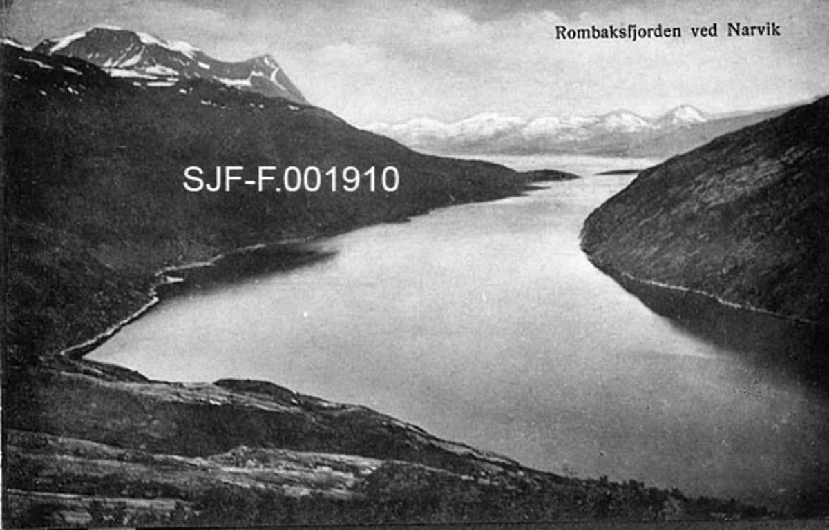 Landskapsbilde fra Narvik kommune i Nordland.  Fotografiet er tatt fra et fjellplatå ned med en fjord, Rombaksbotn, som ligger nordøst for byen.  Fjorden er omgitt av forholdsvis bratte fjell med lite vegetasjon.  På venstre side ser vi Raudbergan.  Til venstre rager Beisfjordstøtta og Rombakstøtta seg over Middagsfjellet. På denne sida skimtes en veg langs strendene.  Bakenfor fjordmunningen skimtes en snødekt fjellkjede på den andre sida av Ofotfjorden. 