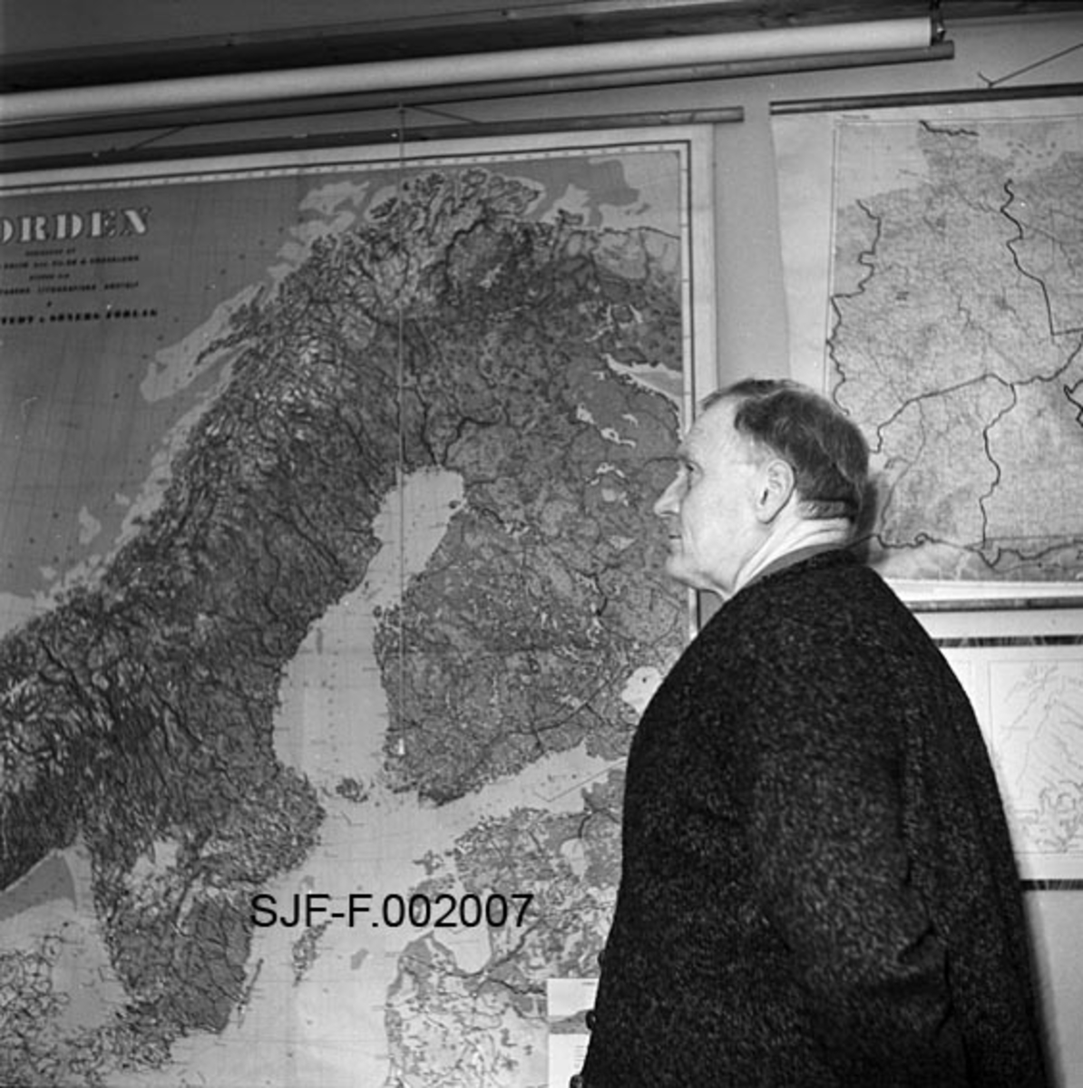 Forstmannen Charles Delgobe Kohmann (1896-1966), fotografert på sine eldre dager, foran et kart over Norden.  Kohmann tok avsluttende eksamen fra skogbruksavdelingen ved Norges landbrukshøgskole i 1919.  Fra 1920 var han fylkesskogassistent i Salten, Lofoten og Vesterålen.  Fra 1922 hadde han oppbygginga og ledelsen av skogplanteskolen i Rognan som hovedoppgave.  Fra 1935 var han dessuten skogbrukslærer ved Nordland landbruksskole.  I 1938 fikk han stillingen som leder for statens planteskole og klenganstalt på Hamar.  Planteskolevirksomheten ble snart flyttet til Åsnes i Solør.  Kohmann fortsatte likevel å lede denne virksomheten fram til 1946.  Da ble klenganstalten på Hamar totalskadd etter en brann, og Kohmann gjorde en kjempejobb, først med å få satt i stand et provisorisk erstatningsanlegg, deretter med å bygge et nytt, stort skogfrøverk som skulle betjene et skogbruk som prioriterte skogreising og planting meget høyt.  Kohmann ledet denne institusjonen inntil han gikk av med pensjon ved årsskiftet 1964-65.  Dette fotografiet ble tatt i 1963. 