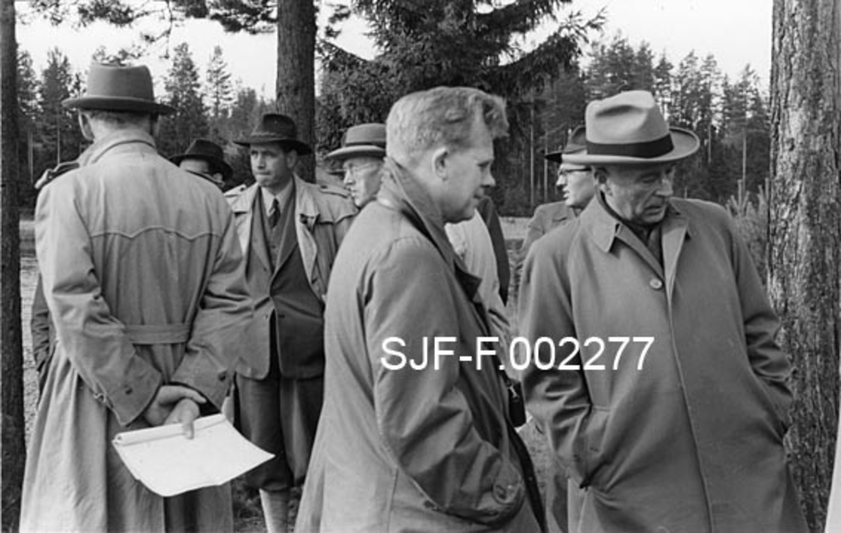 Fra Norsk Fløtingsforbunds ekskursjon ved Flisa-vassdraget 28. september 1954. Fotografiet viser ekskursjonsdeltakerne - antakelig funksjonærer fra fellesfløtingsforeningene - i samtale på et av ekskursjonens stoppesteder.  6-7 menn, de fleste frakkekledde og med hatt på hodet, står og snakker sammen i mindre grupper.  Mannen til venstre i bildet står med ryggen til fotografen, og er følgelig vanskelig å identifisere.  Til høyre for ham står daværende leder for Glomma fellesfløtingsforening, Kaare Sparby (med mørk hatt og frakk som er åpen i fronten, slik at vi ser dressen).  Til høyre for Sparby ser vi hodet til Augustin Paulsen-Næss, som var administrerende direktør i Haldenvassdragets fellesfløtingsforening.  Den barhodete mannen som står like til høyre for bildets midtakse i forgrunnen er sivilingeniør Oddvar Stenerud, som i 1954 var fløtingsinspektør i 7. distrikt med bopæl på Lillehammer.  Han ble seinere administrerende direktør i Glomma fellesfløtingsforening.  Stenerud er fotografert i samtale med forstkandidat Otto Heyner, som var administrerende direktør i Drammensvassdragets fellesfløtningsforening.  Bak nakken (under hattebremmen) til Heyner skimter vi ansiktet til Tore Paulsen-Næss, som den gang var fløtingsinspektør i Glomma fellesfløtingsforening, men seinere ble direktør i Haldenvassdragets fløtningsforening. 

Da disse fotografiet ble tatt hadde Flisavassdraget en femdel av Glommas fløtingskvantum, gjennomsnittlig 225 000 kubikkmeter i året.  Av dette ble cirka 45 000 kubikkmeter fløtet far Halsjøen og tverrelvene i den øvre delen av vassdraget.  Glomma fellesfløtingsforening hadde utført en rekke utbedrings- og forbyggingsarbeider i åra like etter krigen.  Effekten av dette var at fløtinga kunne gjennomføres i løpet av en seksukersperiode, mens en hadde brukt dobbelt så lang tid 50 år tidligere.  Dette var viktig, for Flisavassdraget var fattig på magasiner, og fløtingas effektivitet var derfor i betydelig grad avhengig av vårflommer med begrenset varighet.  "Elveforbedringsarbeidene" gav i tillegg til forkortet fløtingssesong også en økonomisk gunstig tilleggseffekt, nemlig at det hadde vært mulig å redusere bemanninga fra 200-300 til 70-80 mann i perioden mellom 1900 og 1950.  Flisavassdraget ble på 1950-tallet regnet som Glomma fellesfløtingsforenings best utbygde sidevassdrag.