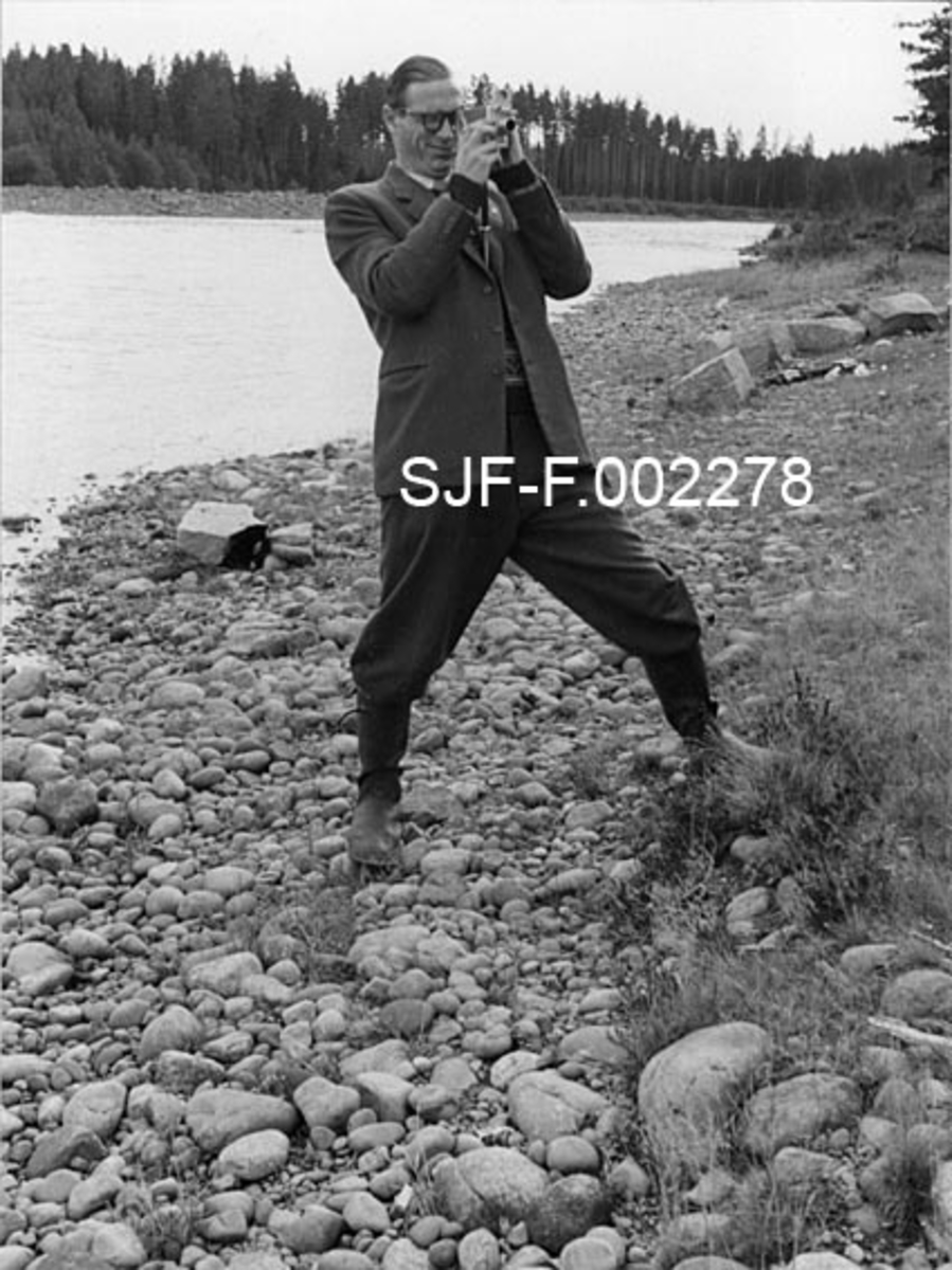 Fra Norsk Fløtingsforbunds ekskursjon ved Flisa-vassdraget 28. september 1954. Fotografiet viser en av ekskursjonsdeltakerne, Sven Gjessing, som på dette tidspunktet var overinspektør i Glomma fellesfløtingsforening.  Han ble seinere adminstrerende direktør i Drammensvassdragets fellesfløtningsforening.  Bildet ble tatt på et tidspunkt da Gjessing fotograferte eller filmet kollegene sine på et av ekskursjonens stoppesteder.  Han er iført vide bukser, langstøvler og dressjakke.  Gjessing har hornbriller og skrått bakoverstrøket hår.  Han står med et kamera hevet foran ansiktet, antakelig vendt mot ekskursjonsdeltakerne.  Bak ham ser vi ei forholdsvis bred, stilleflytende elv med skog langs breddene.  Dette er antakelig Stor-Flisa, muligens ved Kynneggen. 

Da disse fotografiet ble tatt hadde Flisavassdraget en femdel av Glommas fløtingskvantum, gjennomsnittlig 225 000 kubikkmeter i året.  Av dette ble cirka 45 000 kubikkmeter fløtet far Halsjøen og tverrelvene i den øvre delen av vassdraget.  Glomma fellesfløtingsforening hadde utført en rekke utbedrings- og forbyggingsarbeider i åra like etter krigen.  Effekten av dette var at fløtinga kunne gjennomføres i løpet av en seksukersperiode, mens en hadde brukt dobbelt så lang tid 50 år tidligere.  Dette var viktig, for Flisavassdraget var fattig på magasiner, og fløtingas effektivitet var derfor i betydelig grad avhengig av vårflommer med begrenset varighet.  "Elveforbedringsarbeidene" gav i tillegg til forkortet fløtingssesong også en økonomisk gunstig tilleggseffekt, nemlig at det hadde vært mulig å redusere bemanninga fra 200-300 til 70-80 mann i perioden mellom 1900 og 1950.  Flisavassdraget ble på 1950-tallet regnet som Glomma fellesfløtingsforenings best utbygde sidevassdrag. 