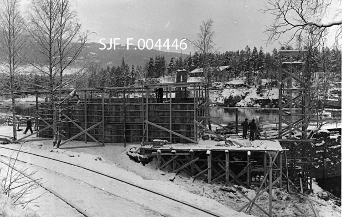 Fra første fase i arbeidet med å bygge en ny dam på toppen av Geithusfossen på Modum i Buskerud.  Dette fotografiet er tatt 14. desember 1961.  I forgrunnen later det til at håndverkere har reist ei slags rampe og noe som minner om et rektangulært skur i forskalingsmaterialer.  Bakenfor skimtes Geithus bru – også kalt «Nybrua» - ei hengebru på fylkesvegen mellom Geithus og Vassbunn.  Den nye dammen ble bygd vinteren etter at Union-konsernet hadde innviet Geithusfoss kraftverk.  Til dette anlegget ble det sprengt en inntakstunnel for vann fra et punkt ovenfor fossen, i fjellet under industriområdet til Drammenselvens Papirfabrikker.  Samtidig ble det også sprengt tømmerrenne utenom fossen.  I desember måned, et par måneder etter at kraftanlegget var ferdig, ble det lagt en fangdam i elvas hovedløp, nedenfor inntaket til kraftstasjonen og ovenfor fossen.  Ved å lede den beskjedne vintervannføringa gjennom de nye avløpene, kunne arbeidet med den nye dammen foregå på noenlunde tørr grunn.  De store arbeidene i elveløpet ble utført i januar, februar og mars 1962, altså etter at dette fotografiet ble tatt. 