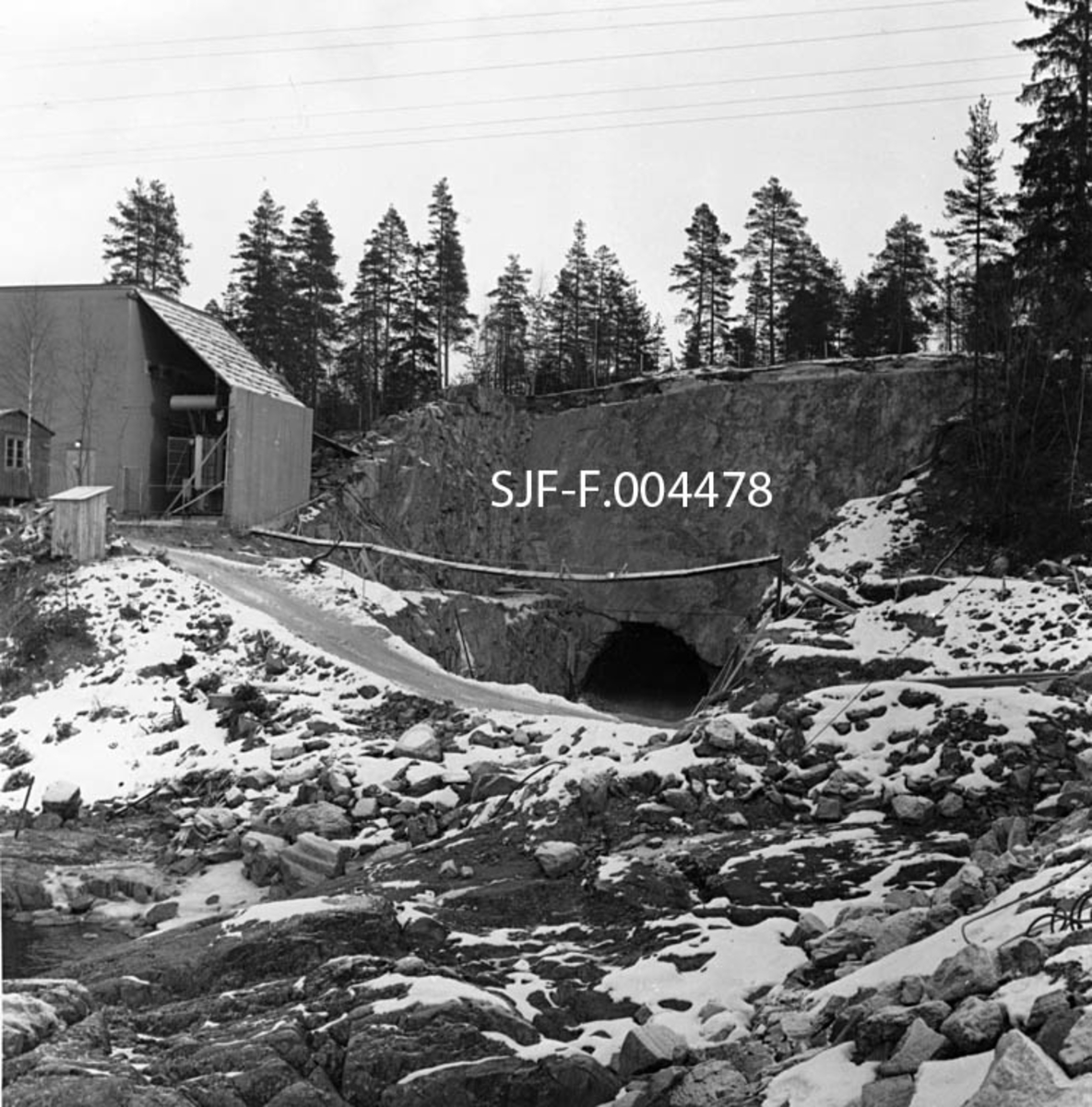 Søndre innslag i driftstunnelen til Geithusfoss kraftverk på Modum i Buskerud.  Fotografiet er tatt fra området der kraftverksbygningen og fordelingsbassenget skulle ligge, mot tunnelen, som i bortimot 110 meters lengde gikk under tomta til Drammenselvens Papirfabrikker.  Dette fotografiet er tatt i januar 1960, et drøyt halvår før gjennomslaget i tunnelen.  Til venstre i bildet sto en murbygning med flatt hovedtak, men med bratt pulttak over et tilbygg mot anleggsområdet.  Vi skimter også gavlen på ei anleggsbrakke, samt en utedo.  En primitiv snødekt gangbru hang over tunnelinnslaget.  På toppen av berget bakenfor vokste det glissen furuskog. 