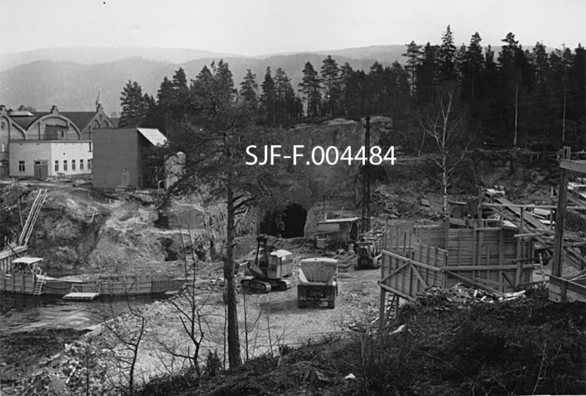 Anleggsområdet der Geithusfoss kraftverk på Modum i Buskerud ble bygd i 1960-61.  Dette fotografiet viser situasjonen i begynnelsen av mai 1960.  Området framfor tunnelen og kraftstasjonstomta var planert for anleggstrafikk.  Da dette fotografiet ble tatt sto det en gravemaskin og en dumper der.  Bakenfor ser vi det søndre innslaget for driftstunnelen i et berg hvor man hadde foretatt en loddrett nedsprengning.  Masse fra tunnelen ble kjørt ut denne vegen.  I forgrunnen ser vi en slags kasseplattform med et elevatorbånd.  Mot vassdraget (nederst til venstre på bildeflata) var det bygd en skjerm av tre.  Til venstre i bildet ser vi noen av bygningene ved Drammenselvens Papirfabrikker.  Det var denne bedriften som hadde fallrettighetene i Geithusfossen, og morselskapet Union Co. finansierte kraftutbygginga blant annet med sikte på å sikre rimelig energi til denne bedriften og andre fabrikker konsernet hadde langs Drammenselva. 