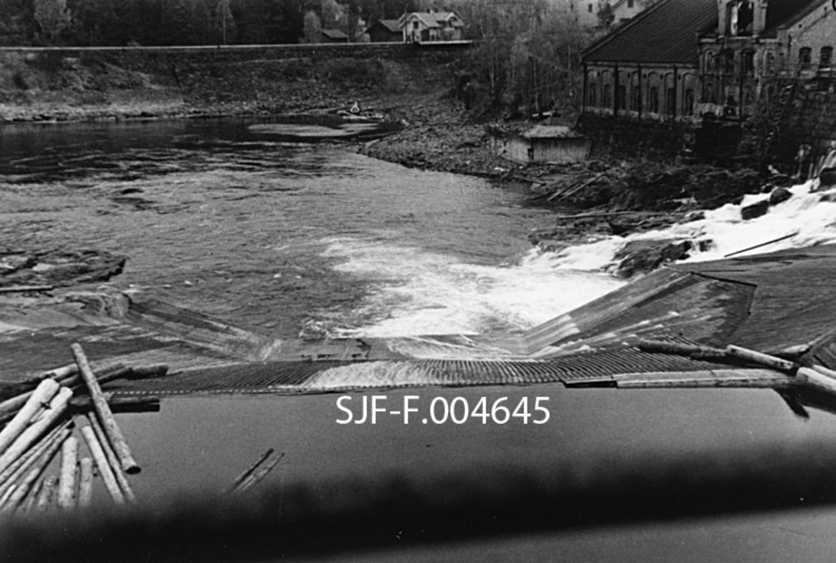 Geithusfossen på Modum i Buskerud, slik den så ut 21. oktober 1945, på et tidspunkt da vannføringa i Drammenselva var ekstremt lav (33 kubikkmeter per sekund).  Fotografiet er tatt i medstrøms retning (akkurat her renner elva nordover) med terskelen som var bygd like ovenfor det øverste fallet i fossen i forgrunnen.  Vi ser hvordan den midtre delen av fallet var golvlagt, med sikte på at tømmer som skulle passere fossen på sin veg videre nedover vassdraget ikke skulle sette seg fast så lett.  Vi ser også enden av ledelensa som skulle bidra til at fløtingstømmeret ble ledet mot nettopp denne golvlagte delen av fallet.  Ved elvebredden til høyre i bildet ligger tresliperiet som leverte tremasse til Drammenselvens Papirfabrikker, på den motsatte sida av fossen.  Tresliperiet var et teglsteinsbygg i tidstypisk industriarkitektur.  Bygningen ble herjet av brann noen år etter at dette fotografiet ble tatt, og aldri gjenoppført.  Sentralt i bakgrunnen ser vi Hontvedts forretning.  Vi skimter også (bak og til venstre for taket på sliperibygningen) litt av Modums samvirkelags bygg med bakeri, pølsemakeri, lager og kontorer for administrasjonen. 