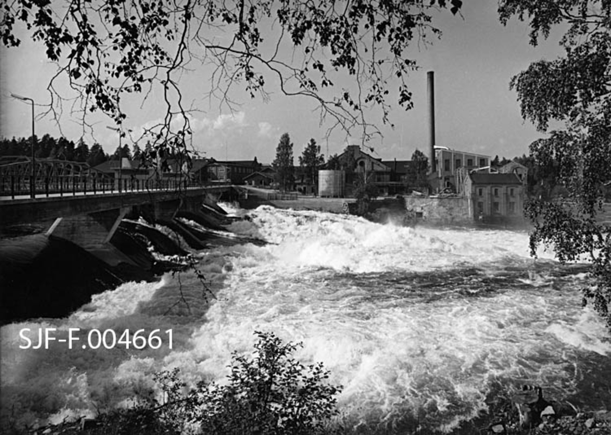 Det øverste fallet i Geithusfossen på Modum i Buskerud.  Fotografiet er antakelig tatt forholdsvis kort tid etter at Union-konsernet vinteren 1961-62 hadde bygd ny dam er, blant annet for å kunne heve vannspeilet ved det ovenforliggende inntaket til driftstunnelen til Geihusfos kraftverk (1960-61) og til den nye tømmerrenna, som også var innsprengt i berget vest for Drammenselvens Papirfabrikker.  Det er noe av bebyggelsen ved denne bedriften vi ser på motsatt side av elva her.  Bildet er tatt på et tidspunkt da elva var stor og strid, antakelig i forbindelse med vårflommen i mai-juni.  Fotografen har stått på vestre elvebredd, under et bjørketre, med greiner som «rammer inn» fossen.  Til venstre i bildet ser vi den nye betongdammen med brubane og «gatebelysning».  På motsatt side av elva ser vi industribebyggelsen ved Drammenselvens Papirfabrikker. 