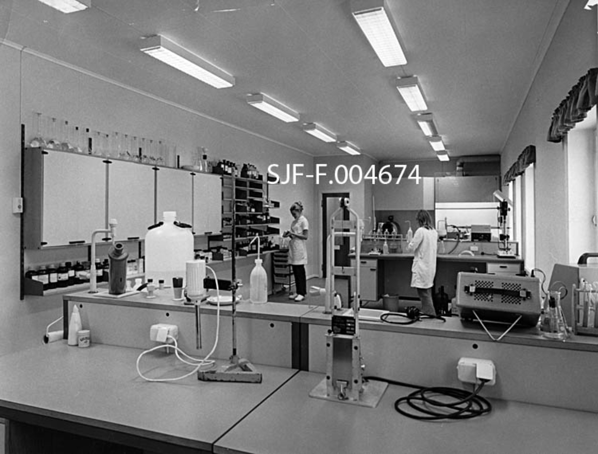 Fra laboratoriet ved Embretsfoss fabrikker på Åmot i Modum kommune i Buskerud.  Fotografiet er antakelig tatt i begynnelsen av 1970-åra.  Vi ser et rektangulært rom med kvitmalte vegger og tak.  Rommet lyses opp av dagslys som faller inn fra vinduer på langveggen og fra to rekker av rektangulære lamper med lysrør under gitter.  Møblementet består av tverrstilte arbeidsbenker med diverse laboratorieutstyr.  På den vinduløse langveggen til venstre i bildet er det skap og hyller med kjemikalieflasker og reagensglass.  I enden av rommet aner vi et avtrekkskap.  Vi ser også to laboranter, to unge kvinner i kvite frakker. 