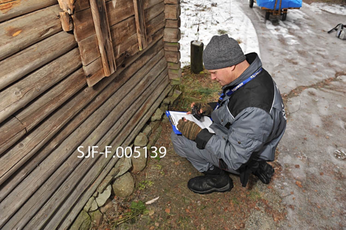 Tømrer Knut-Arild Nordli undersøker en skogsstall fra Sørskogbygda i Elverum [SJF-B. 0014] i Norsk Skogmuseums friluftsmuseum på Prestøya.  Nordli sitter på huk på nordsida av bygningen, under et takoverbygg over to luker i ytterveggen, som gjorde det mulig å fôre hestene uten å gå inn i stallrommet.  Hani gjør notater på et medbrakt skriveunderlag.  Tømreren er kledd i grå arbeidsdress med svarte skulderstykker og har grå lue på hodet.  Våren 2012 ble Nordli engasjert for å gjennomgå de antikvariske bygningene i Hedmark fylkesmuseum med sikte på å definere materialbehov og tidsforbruk ved restaureringstiltak ved fylkesmuseets åtte avdelinger.  Vurderingsarbeidet i Skogmuseets friluftsmuseum på Prestøya ble utført seinhøstes 2012. 