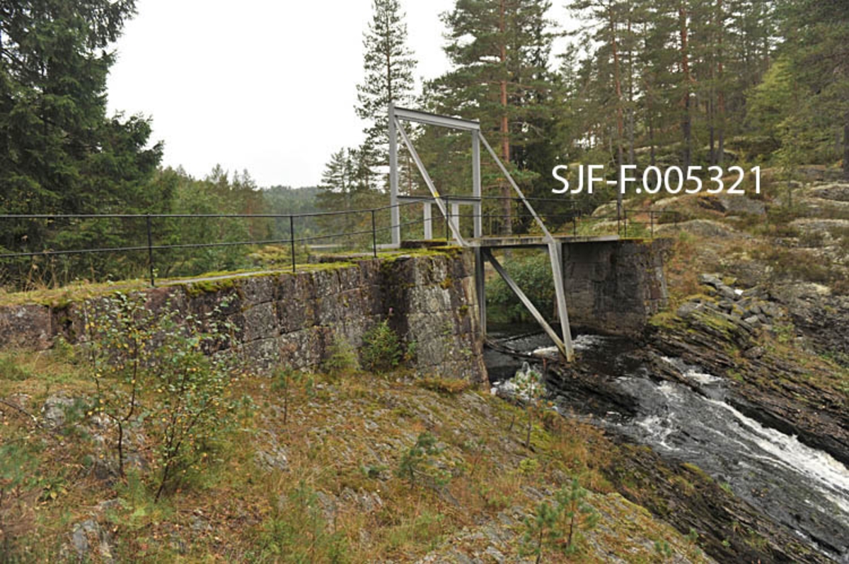 Vigdesjådammen ved utløpet av tjernet Vigdesjå i Kviteseid i Vest-Telemark.  Dammen er bygd av hoggen naturstein med krone av betong.  Dambrua har jernrekkverk på medstrøms side og ei stålramme for damlukene på motstrøms side.  Her ved utløpet av Vigdesjå renner Bygdaråi øst- og etter hvert nordøstover i bergrikt lende med Damåsan på nordsida og Kolakosi på sørsida, mot Høgfossen og Kolbjørnsrudfossen,  Fotografiet viser dammen omgitt av et landskap med blandingsskog (mye furu) og bergflater med lav- og lyngvegetasjon.  Bildet er tatt i vestlig retning mot damkonstruksjonen og damåpningen, hvor det er et visst fall i terrenget. 