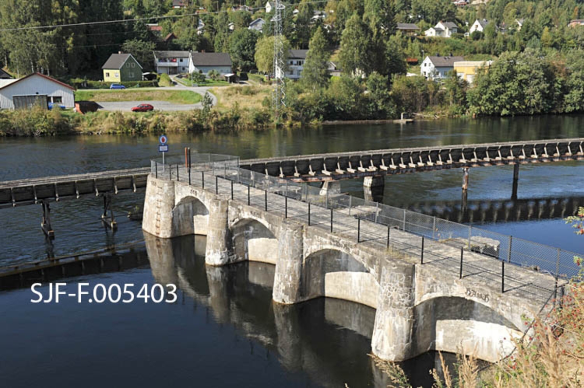 Fra den såkalte Sagafossdammen på Notodden, der Tinne (Tinnelva) ble stengt med en dam, som var vannreservoar for kraftstasjonene Tinfos I og Tinfos II.  Kraftstasjonen Tinfos I sto ferdig i 1901.  Dette kraftverket gav kraft til Holtas bedrifter, en treforedlingsfabrikk og en karbidfabrikk.  I bakken øst for Sagafoss planla Holta en 900 meter lang kanal som skulle bli vannmagasin for det planlagte nye kraftverket, Tinfos II.  Vest for kanalen ble det lagt en jordvoll mot dalsida. Etter planen skulle slam fra elva tette denne vollen, noe som viste seg å ta tid.  Det nye kraftverket ble satt i drift i 1912.  Fallhøyden fra kanalen var på 28, 5 meter, og de tre turbinene gav mye energi, blant annet til Tinfos Jernverk, hvor den samme Holta også var sentral som eier.  De mange kraftverksprosjektene i Tinne/Tinnelva gjorde det nødvendig å bygge tømmerrenner som skulle ta tømmeret forbi kraftverksdammene.  I 1920-åra ble det bygd ei renne fra Lienfossdammen, forbi Sagafoss og Tinnfoss.  Denne renna ble 1 863 meter lang.  Den nedre delen av den renna står på søyler i dammen på dette fotografiet.  Fløtinga på Tinnsjøen ovenfor opphørte i 1970.  Seinere har det bare unntaksvis gått tømmer i renna, siste gang i 1992.  På denne tida innså man at renna var et viktig kulturminne.  I midten av 1990-åra ble det igangsatt restaureringstiltak i samarbeid med kommunale og regionale verneinteresser og Skiensvassdragets Fellesfløtningsforening, som fridde seg fra videre ansvar ved å skyte inn et beløp som tilsvarte stipulerte rivingskostnader.  I 2012-13 vurderer Riksantikvaren fredning av tømmerrenna på Notodden.