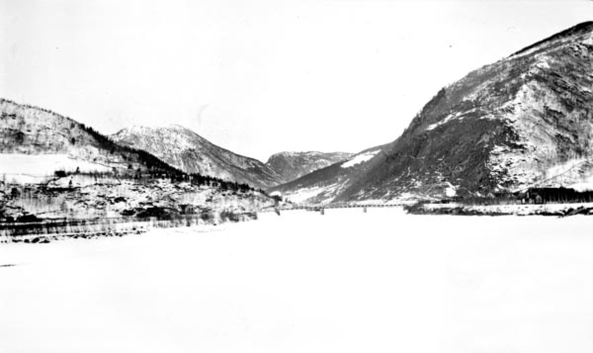 Vinterlandskap ved tettstedet Otta i Sel kommune i Gudbrandsdalen.  Fotografiet later til å være tatt fra den snødekte isflata på elva Lågen.  Sentralt i bildet ser vi ei fagverksbru av stål i tre spenn mellom steinkar i elveløpet.  På sletta til høyre skimtes noe bebyggelse, antakelig det nevnte tettstedet.  Ellers preges landskapet av til dels bratte fjell med glissen skogkledning. 