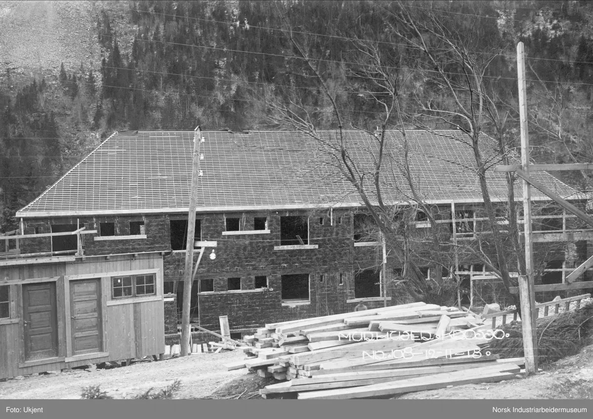 Murhusbebyggelsen på Krosso under oppføring. Bygget har fått vegger og det er lagt sløyfer og lekter på taket. Vinduer og dører mangler. Brakke på veien ovenfor bygget. Stabler med materialer liggende ved veien. Gatebelysning i strømstolpe.