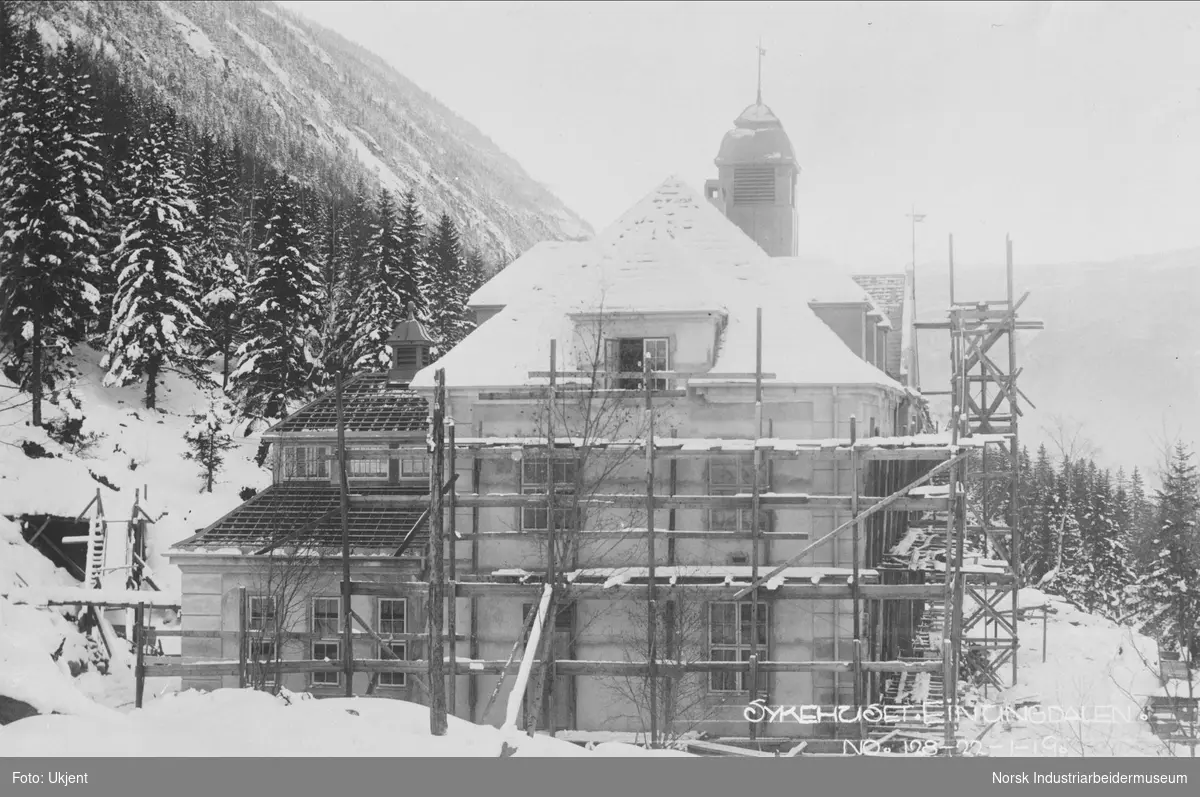 Bygging av sykehuset i Einungdalen. Veggene er ferdig reist, taket er i ferd med å legges. Trestillas stående rundt bygningen.