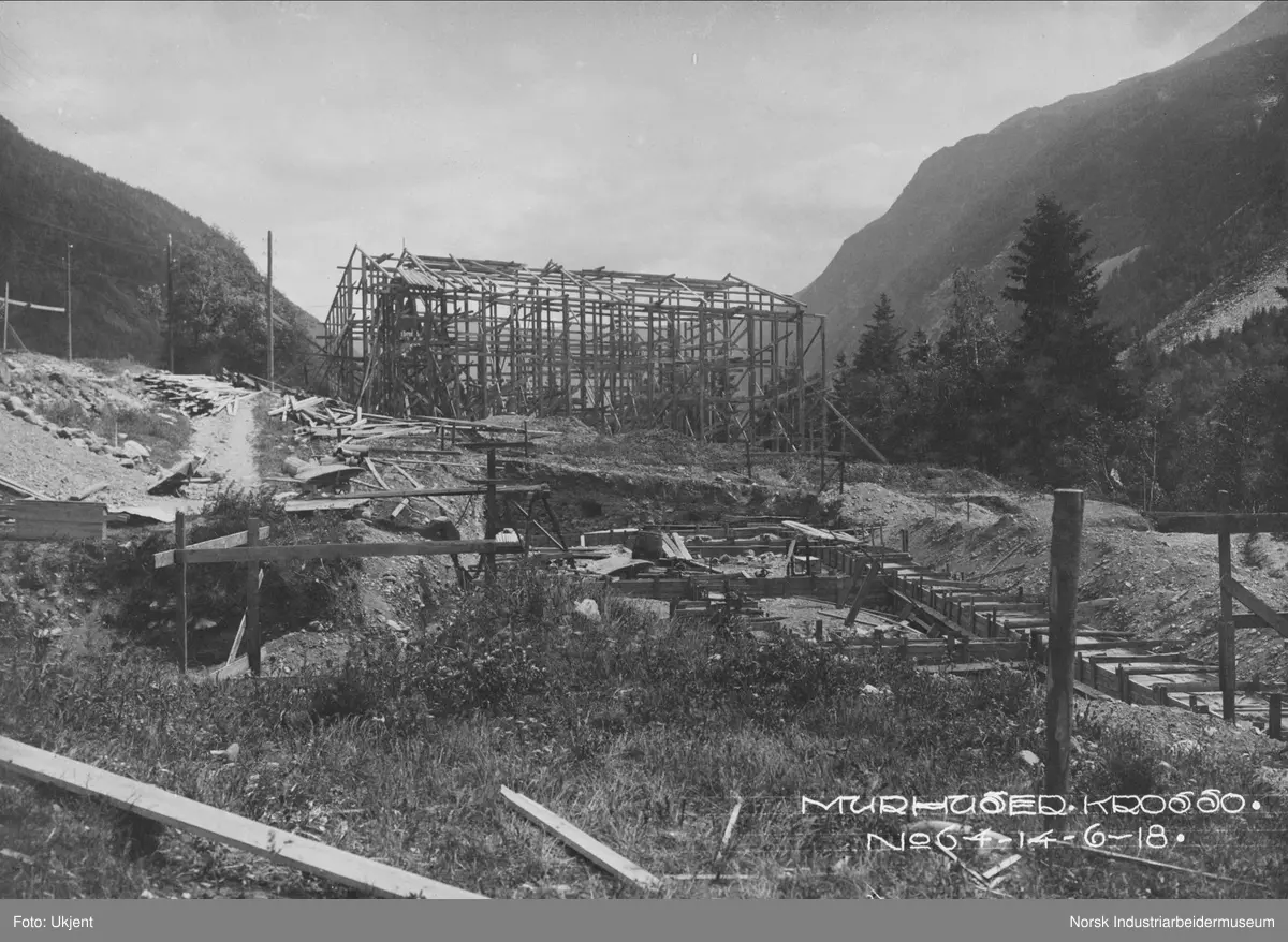 Murhusbebyggelsen på Krosso under oppføring. Grunnarbeid for murhus, grunnmur er støpt. Reisverk til bygg i tre i bakgrunnen.