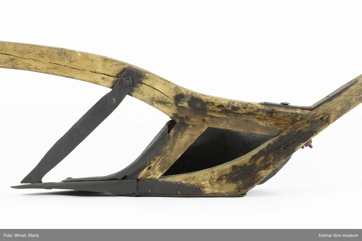 KLM 12823. Plog, trä, metall. Plog med handtag och ås av trä. På åsens ena sida sitter en rist av järn fastskruvad. Längst ut på åsen en dragkrok av järn för anspänning av dragdjur. Plogblad med bill av järn. Träkil mellan plogblad och landsida. Plogen är järnskodd på undersidan. Den har även varit järnskodd på landsidan men denna saknas. Spikarna sitter kvar i träet. Fyra slåar mellan handtagen, två av järn och två av trä. Det nedersta är fastskruvat i plogbladet. Plogen är svartmålad med en röd eventuell rostskyddsfärg under. Målad under senare tid.