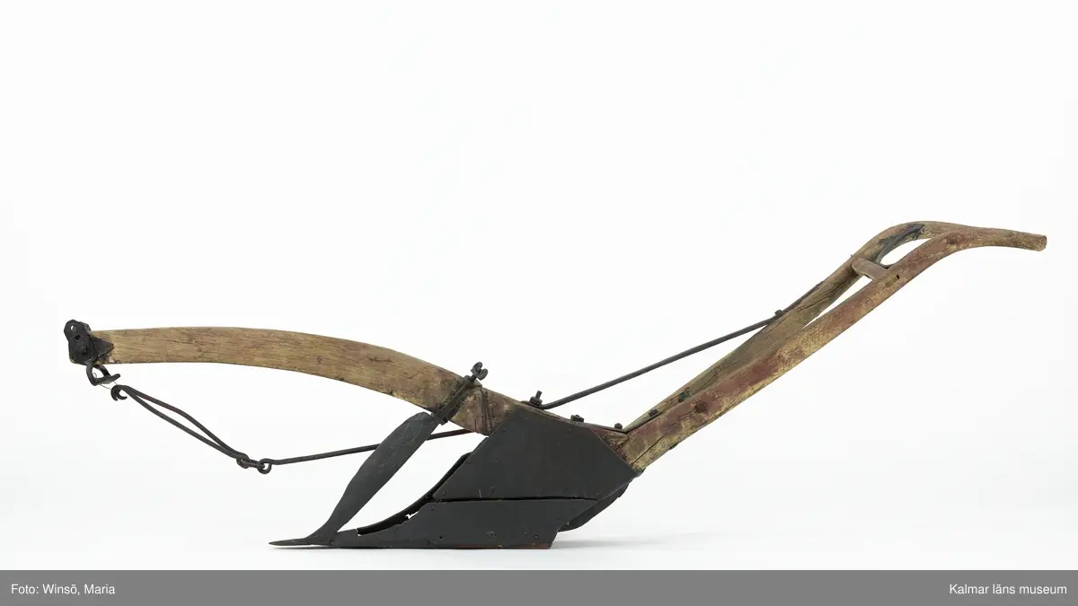 KLM 12824. Plog, trä, metall. Plog med handtag och ås av trä. Landsida av trä, järnskodd. Vändskiva och bill av järn. Tvärslå av järn mellan handtag och ås. Slåar mellan handtagen, två rundformade av järn, en U-formad av järn, en kantig av järn och en av trä. Längst fram på åsen en dragkrok av järn med reglage för inställning av höjd. På åsens mitt en fastskruvad rist. Risten är sammanfogad, eventuellt med färg, med billen. Tre långa krokar av järn hänger under åsen och möts i en järnring. Plogen är trasig på landsidan, träet är sprucket och järnbeslaget har lossnat. Plogen är rödmålad men färgen är kraftigt avskavd. Plogens järndetaljer är svartmålade med en röd eventuell rostskyddsfärg under. Den svarta färgen och rostskyddsfärgen är målad senare.