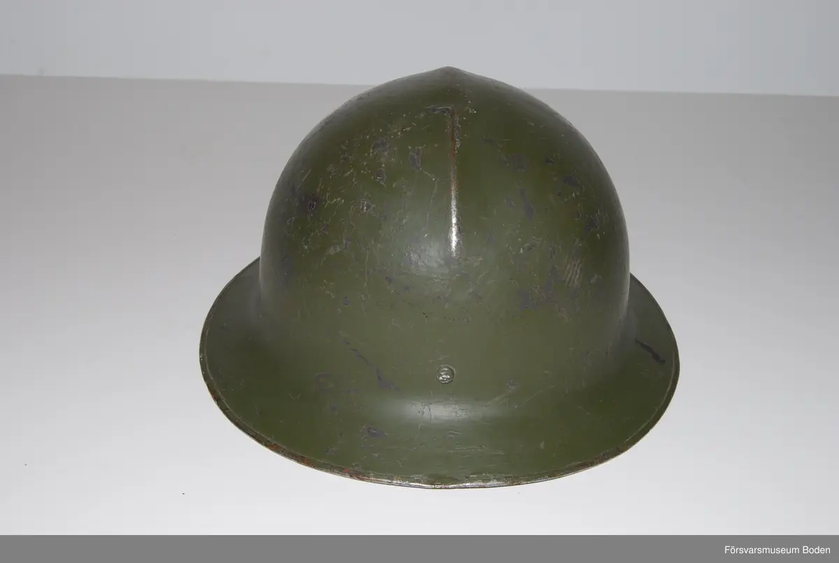 Olivgrön stålhjälm av typen med bredare och plattare brätte, den till vänster på bild 6 som jämför båda typerna av hjälm m/1921. Med insats bestående av tre läderkuddar som hålls ihop med skorem. Hakband av läder. Framtill tre kronor i relief, inga dekaler på sidorna. Upptill en förstärkningspressning i form av en kam.