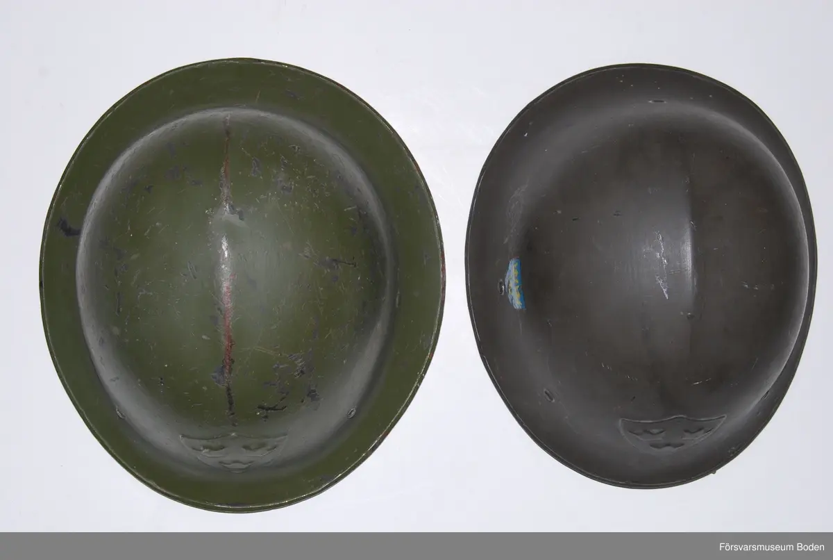 Olivgrön stålhjälm av typen med bredare och plattare brätte, den till vänster på bild 6 som jämför båda typerna av hjälm m/1921. Med insats bestående av tre läderkuddar som hålls ihop med skorem. Hakband av läder. Framtill tre kronor i relief, inga dekaler på sidorna. Upptill en förstärkningspressning i form av en kam.