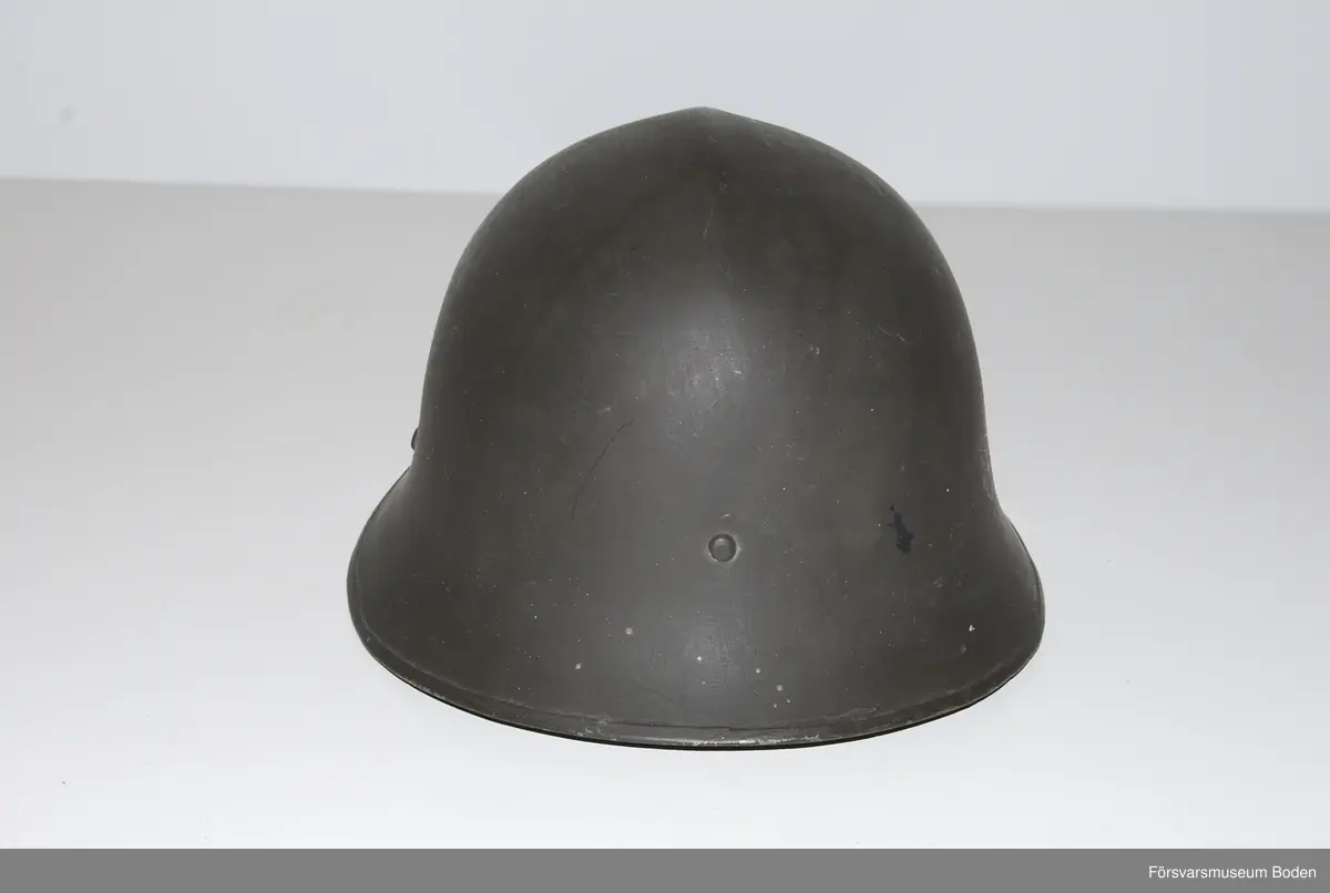 Mörkt grågrön stålhjälm av typen med smalare och högre brätte, den till höger på bild 6 som jämför båda typerna av hjälm m/1921. Med insats bestående av tre läderkuddar som hålls ihop med skorem. Hakband av läder. Framtill tre kronor i relief, tre-kronor dekaler på sidorna. Upptill en förstärkningspressning i form av en kam.