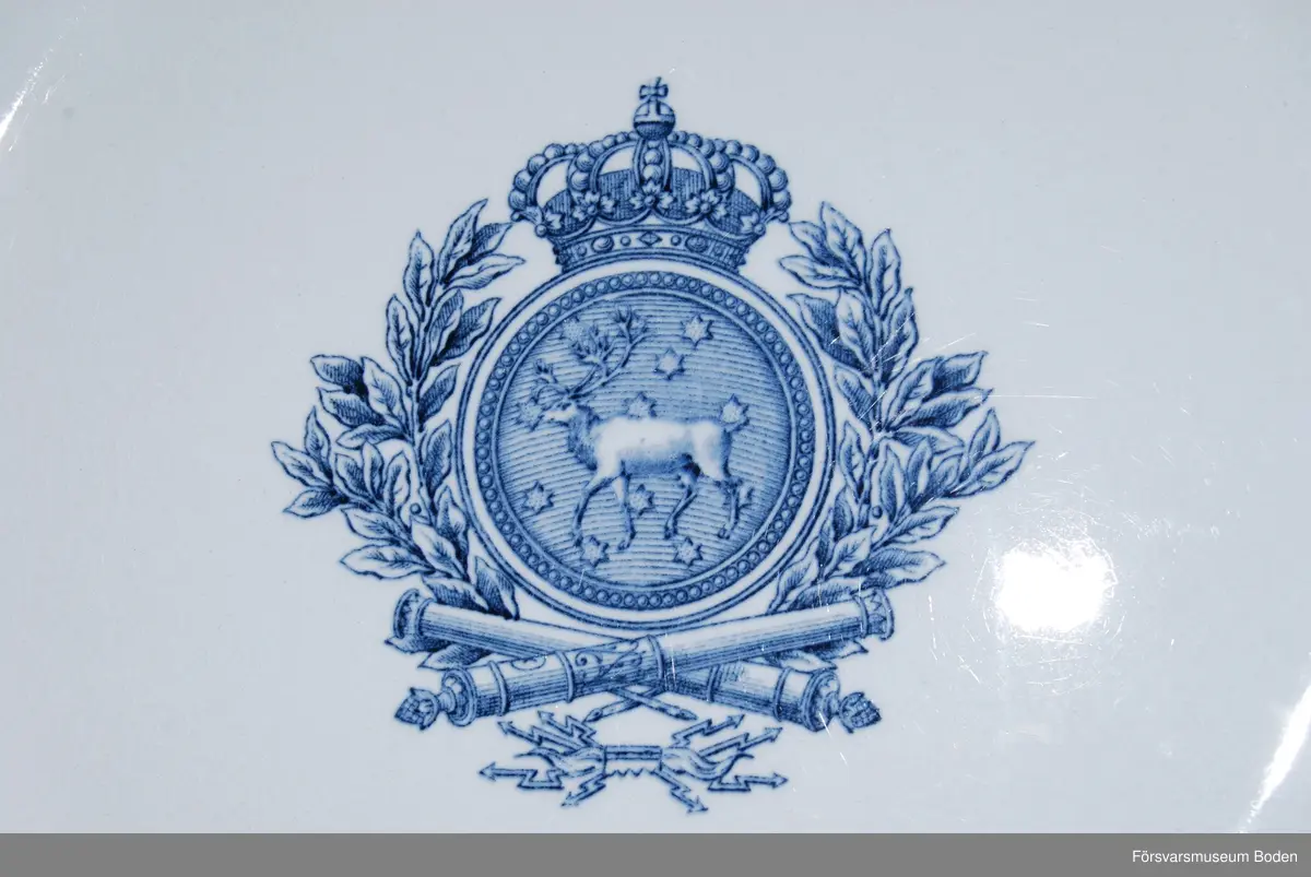 Flat 8-kantig tallrik av blåtonat porslin (flintgods) av Rörstrands tillverkning. I centrum en blå vapenbild med Västerbottens landskapsbild i rund, krönt sköld omgiven av lagerkrans. Nertill under skölden korslagda eldrör och knippe med blixtar.