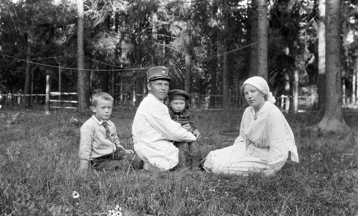 En soldat (offiser/befal), en kvinne og to barn som sitter på grasbakken i parklignende omgivelser. Identitet ukjent. Sted trolig Gardermoen.