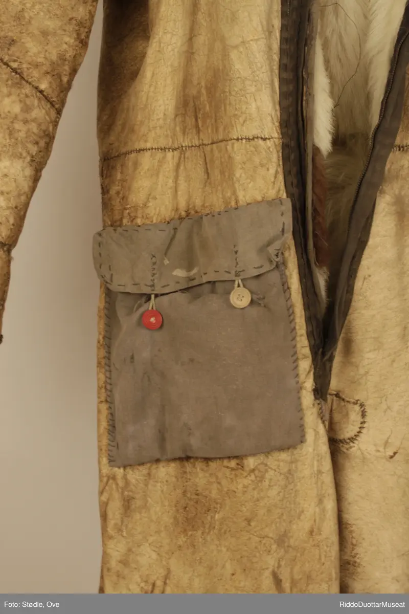 Hel dress, 173 lang. Sydd av saueskinn og sannsynligvis storfeskinn, med hårsiden innover.  Kragen er sydd av saueskinn med ullsiden ut. Glidelås-lukning. Ligner en gammel flydress. Lomme øverst på høyre lår er sydd av grått stoff, og har en rød og en brun knapp.