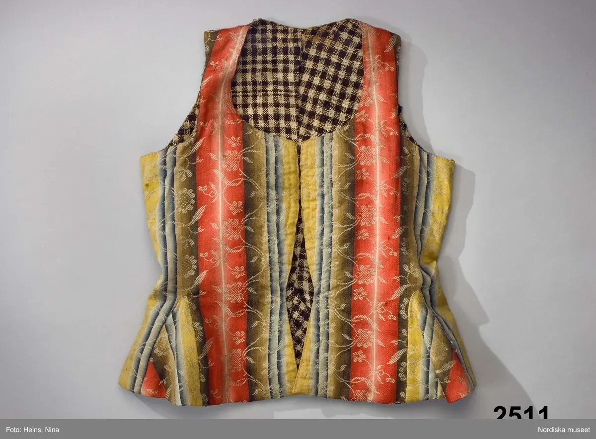 Livstycke med långt skört, av finaste kamgarnsylle i satin, randigt i varpled i breda bårder i tonande nyanser av rött, grönt, gult och blått med invävda blomsterslingor i vitt som ligger på en extra mönstervarp. Tygkvaliten kallas blommerad kalmink och har från början varit starkt glättat. 1700-talsmodell med 2 framstycken/sidstycken med skörtveck i den bakåtlagda sidsömmen och infälld kil i framstycket. Framkanterna något svängda. 2 ryggstycken med skörtveck i baksömmen. Dold knäppning fram med 4 par hakar och hyskor av järntråd. Foder av hemvävt rutigt linne i gles tuskaft, rutigt i mörkbrunt och ofärgat lingarn.
Anm. Något solkigt, på ena bakstycket en stoppning i midjelinjen. Nedersta haken saknas.
Kalmink blev modernt i slutet av 1700-talet till högtidsdräkter främst i bröllopsplagg. Det var ett köpetyg som kom från England där Norwich vävde de allra finaste kvaliteterna. Förmodligen inkommet via gränshandel med Norge.
/Berit Eldvik 2007-06-11