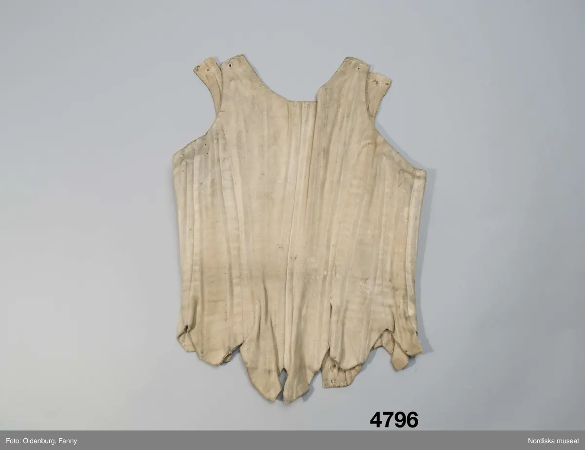 Snörliv av ljust sämskskinn. Skuret i 10 smala delar med 20 kanaler för styvnader av valben. Axelbanden öppna med ett snörhål i var ände för reglering av längden medelst knytband. I framkanterna snörning med ursprungligen 8 par snörhål kantsydda med lintråd. Helfodrat med oblekt linnelärft.
Har förändrats för att bli mindre. Dels har minskningen gjorts mitt bak med en ny söm, dels har framkanterna vikts in och sytt fast mot fodret och 8 par nya snörhål har prylats och kantsytts. På framstyckena finns även sekundärt gjorda insyningar som sedan sprättats upp.
Snörliv med valben var dyrbara och kan ha gått i arv från t.ex. mor till dotter och då sytts om för att bli smalare.
Anm. Solkigt och bitvis slitet invändigt med hål vid kanalerna där valben syns genom hålen. Flera valbenen saknas också. Troligen har det från början varit järntenar i framkanterna vid den ursprungliga snörningen.
Har enl. förmedlaren tillhört prostinnan Sara Hazelius i Bergsjö, Hälsingland. Sådana snörliv bars inte av allmogekvinnorna.
Sara f. Eneroth 28/9 1726 var dotter till kyrkoherde Pehr Eneroth i Svenljunga, Älvsborg. Hon gifte sig 1756 med sedemera prosten i Bergsjö Pehr Hazelius och fick 8 barn 1757-1768. Hon dog 1795.
Läs om snörliv B.Hammar/P.Rasmussen: Underkläder, Signum förlag 2008 
/Berit Eldvik 2008-05-21