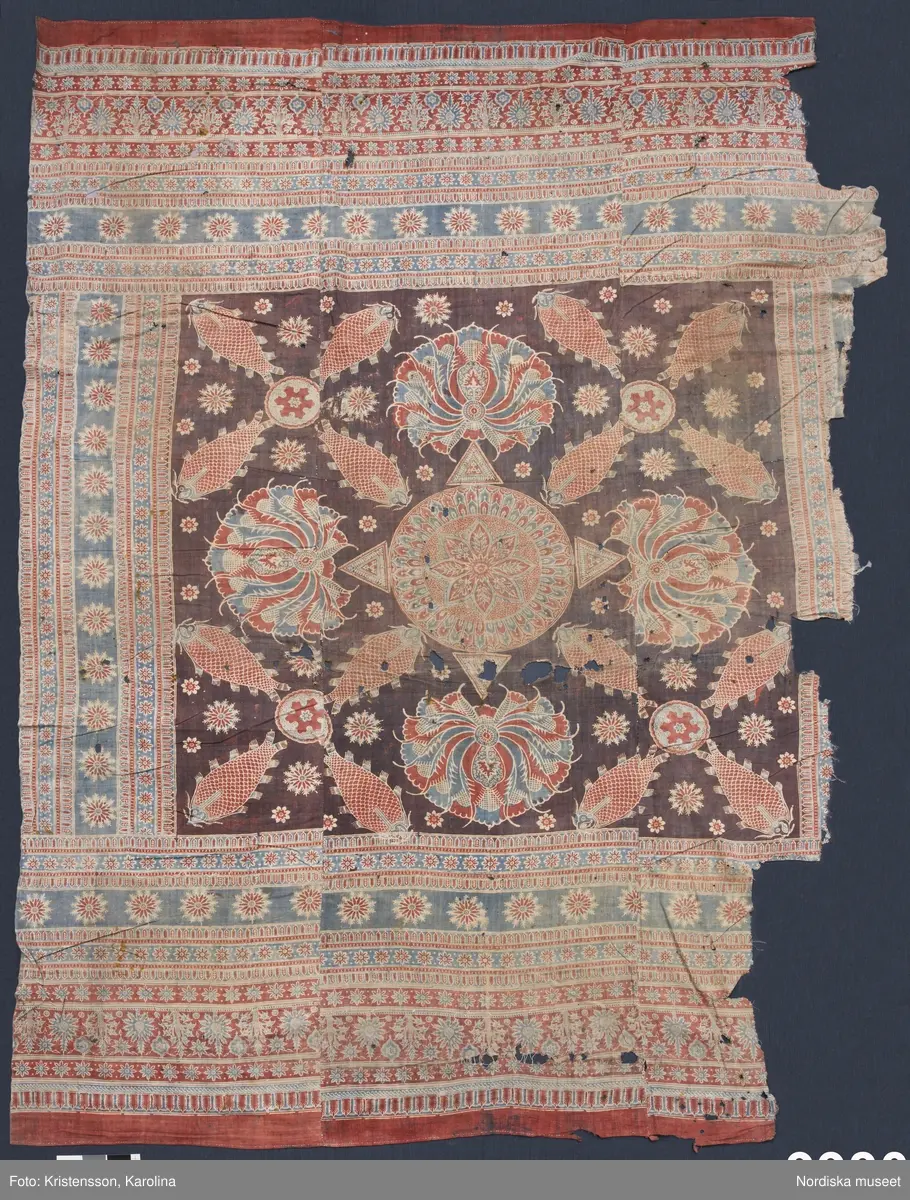 Stort tygstycke sammansytt av 3 våder bomullslärft  á 54 cm bredd. Hopsydda med vitt handspunnet lingarn. Tryckt mönster i rött och blått på vitt underlag.. I mitten stort cirkelmotiv fylld med mönster och i mitten en stjärna, åt fyra håll en triangel ut från cirkeln och mot 4 större snäckliknande ornament. I varje hörn motiv med 4 fiskar med stjärtarna in mot en cirkel, Kantbårder bestånde av olikmönstrade ränder i olika bredder.  Trycket är gjort med tryckstockar och smalare tryckstämplar i kantbårderna där tydliga blockskarvar syns.  Tekniken är den gamla indiska med betmålning av de partier som skall bli röda och därefter doppning i krappbad, tvättning av tyget före blåfärgning som görs så att vita och röda parteier täcks med en pasta före färgning i indigokyp. På baksidan är tyget mycket blåare än på framsidan. Spegelns botten har färgats först röd och därefter blå  så att den fått en mörklila , lätt flammig nyans. Tyget är skört och trasigt på flera ställen, längs hela högersidan är det söndigt vilket kan vara musätet. En del mindre hål har lappats med  gråblått siden på baksidan eller på ett ställe med en bit av själva tygtrycket. Även blekt.
Insamlat av Nils Månsson-Mandelgren vid hans resor i Västergötland, inga uppgifter finns.
 /Berit Eldvik 2013-06