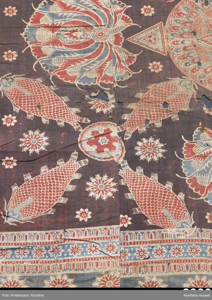 Stort tygstycke sammansytt av 3 våder bomullslärft  á 54 cm bredd. Hopsydda med vitt handspunnet lingarn. Tryckt mönster i rött och blått på vitt underlag.. I mitten stort cirkelmotiv fylld med mönster och i mitten en stjärna, åt fyra håll en triangel ut från cirkeln och mot 4 större snäckliknande ornament. I varje hörn motiv med 4 fiskar med stjärtarna in mot en cirkel, Kantbårder bestånde av olikmönstrade ränder i olika bredder.  Trycket är gjort med tryckstockar och smalare tryckstämplar i kantbårderna där tydliga blockskarvar syns.  Tekniken är den gamla indiska med betmålning av de partier som skall bli röda och därefter doppning i krappbad, tvättning av tyget före blåfärgning som görs så att vita och röda parteier täcks med en pasta före färgning i indigokyp. På baksidan är tyget mycket blåare än på framsidan. Spegelns botten har färgats först röd och därefter blå  så att den fått en mörklila , lätt flammig nyans. Tyget är skört och trasigt på flera ställen, längs hela högersidan är det söndigt vilket kan vara musätet. En del mindre hål har lappats med  gråblått siden på baksidan eller på ett ställe med en bit av själva tygtrycket. Även blekt.
Insamlat av Nils Månsson-Mandelgren vid hans resor i Västergötland, inga uppgifter finns.
 /Berit Eldvik 2013-06