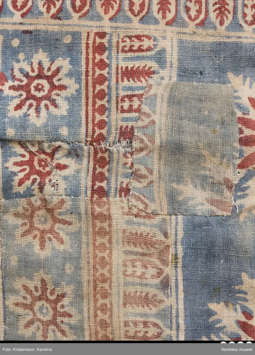 Stort tygstycke sammansytt av 3 våder bomullslärft  á 54 cm bredd. Hopsydda med vitt handspunnet lingarn. Tryckt mönster i rött och blått på vitt underlag.. I mitten stort cirkelmotiv fylld med mönster och i mitten en stjärna, åt fyra håll en triangel ut från cirkeln och mot 4 större snäckliknande ornament. I varje hörn motiv med 4 fiskar med stjärtarna in mot en cirkel, Kantbårder bestånde av olikmönstrade ränder i olika bredder.  Trycket är gjort med tryckstockar och smalare tryckstämplar i kantbårderna där tydliga blockskarvar syns.  Tekniken är den gamla indiska med betmålning av de partier som skall bli röda och därefter doppning i krappbad, tvättning av tyget före blåfärgning som görs så att vita och röda parteier täcks med en pasta före färgning i indigokyp. På baksidan är tyget mycket blåare än på framsidan. Spegelns botten har färgats först röd och därefter blå  så att den fått en mörklila , lätt flammig nyans. Tyget är skört och trasigt på flera ställen, längs hela högersidan är det söndigt vilket kan vara musätet. En del mindre hål har lappats med  gråblått siden på baksidan eller på ett ställe med en bit av själva tygtrycket. Även blekt.
Insamlat av Nils Månsson-Mandelgren vid hans resor i Västergötland, inga uppgifter finns.
 /Berit Eldvik 2013-06