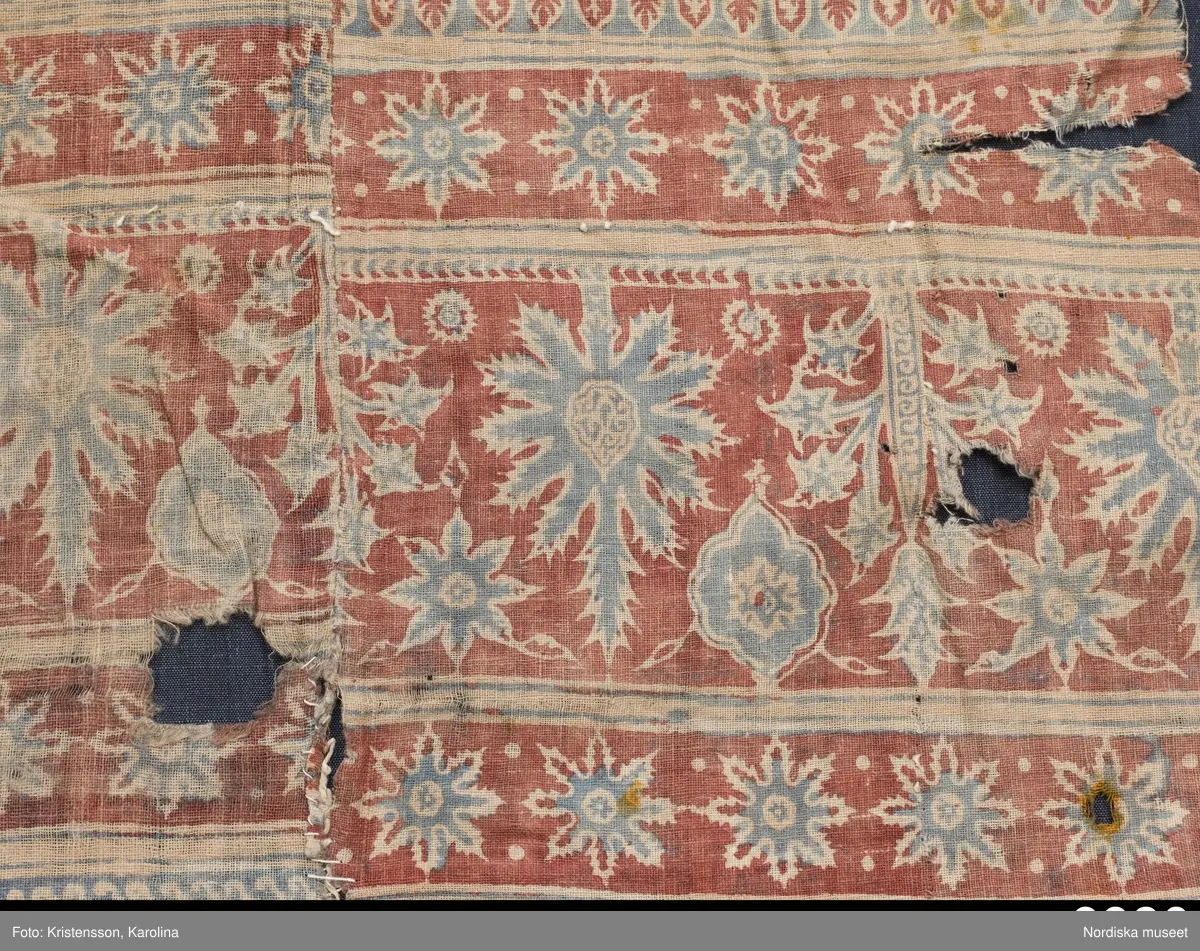 Stort tygstycke sammansytt av 3 våder bomullslärft  á 54 cm bredd. Hopsydda med vitt handspunnet lingarn. Tryckt mönster i rött och blått på vitt underlag.. I mitten stort cirkelmotiv fylld med mönster och i mitten en stjärna, åt fyra håll en triangel ut från cirkeln och mot 4 större snäckliknande ornament. I varje hörn motiv med 4 fiskar med stjärtarna in mot en cirkel, Kantbårder bestånde av olikmönstrade ränder i olika bredder.  Trycket är gjort med tryckstockar och smalare tryckstämplar i kantbårderna där tydliga blockskarvar syns.  Tekniken är den gamla indiska med betmålning av de partier som skall bli röda och därefter doppning i krappbad, tvättning av tyget före blåfärgning som görs så att vita och röda parteier täcks med en pasta före färgning i indigokyp. På baksidan är tyget mycket blåare än på framsidan. Spegelns botten har färgats först röd och därefter blå  så att den fått en mörklila , lätt flammig nyans. Tyget är skört och trasigt på flera ställen, längs hela högersidan är det söndigt vilket kan vara musätet. En del mindre hål har lappats med  gråblått siden på baksidan eller på ett ställe med en bit av själva tygtrycket. Även blekt.
Insamlat av Nils Månsson-Mandelgren vid hans resor i Västergötland, inga uppgifter finns.
 /Berit Eldvik 2013-06