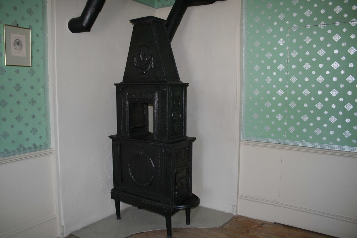 Gammel jernovn, Louis XVI stil.
2 etasjer m/pyramidetopp, kvinnefigur m/brennende alter i medaljong vase og krans.  Rosett i hjørner, står Eidsvold i medaljong, og CA på ovnsdøra.