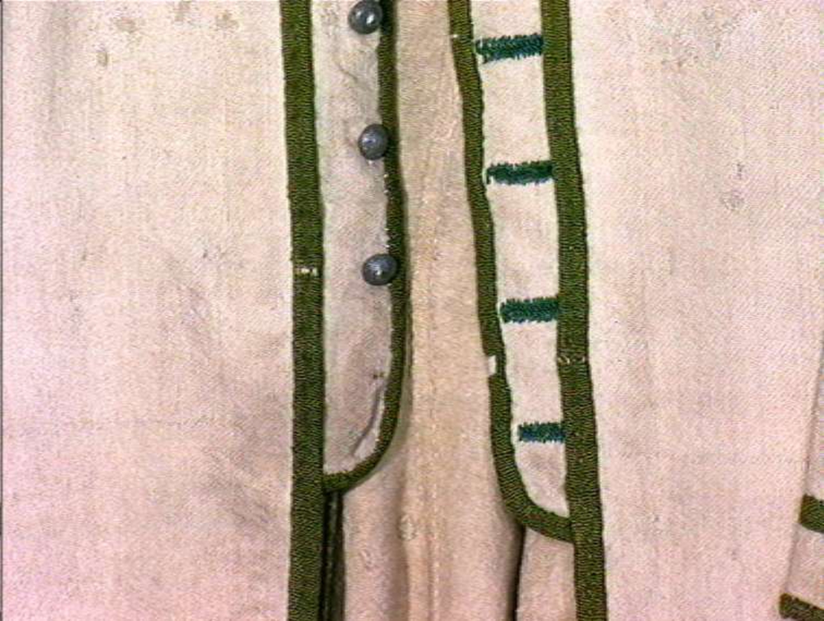 Hvit kjole av ull med grøn dekor av ull.
Midjen bak er markert med to garn dusker.