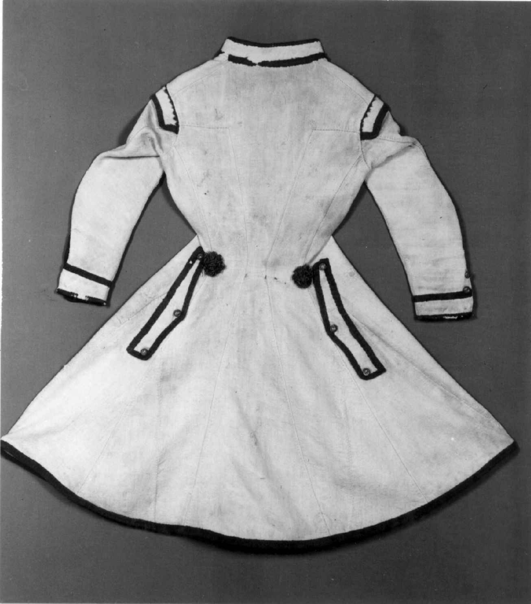 Hvit kjole av ull med grøn dekor av ull.
Midjen bak er markert med to garn dusker.