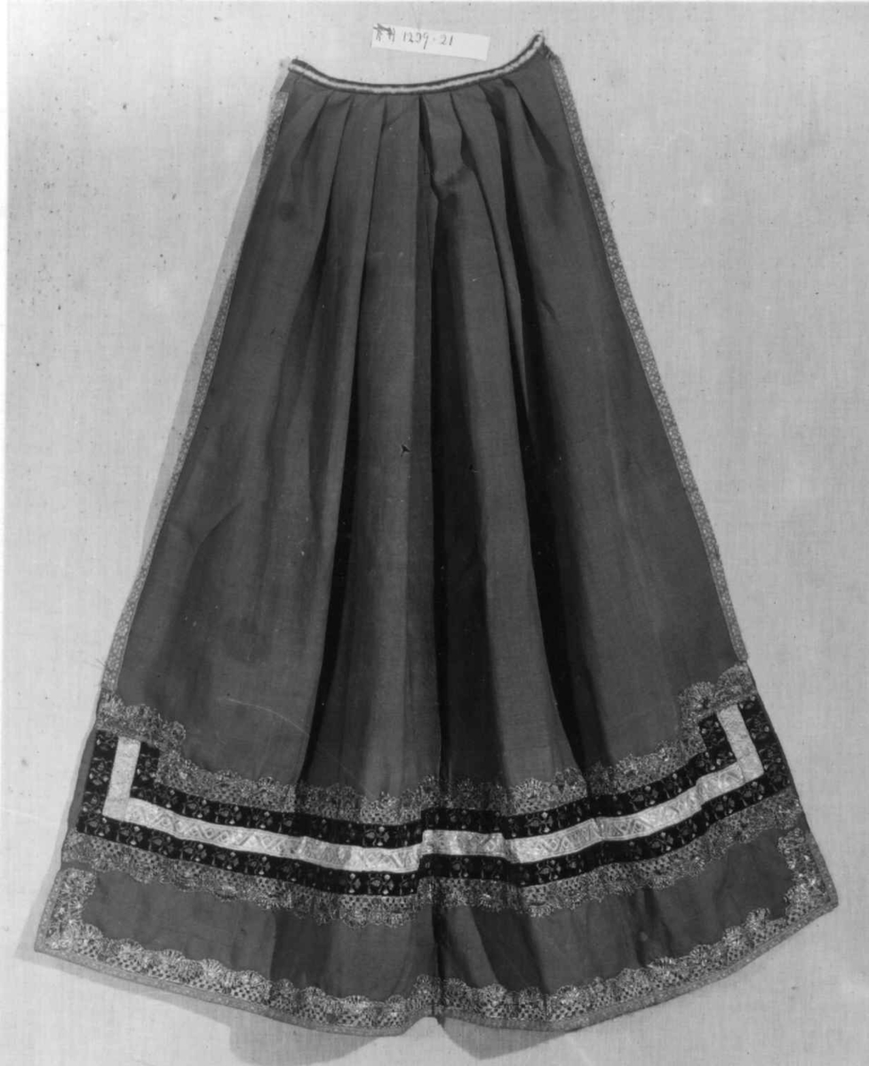 Grønt forkle av ull med dekor av metallblonder og bånd.