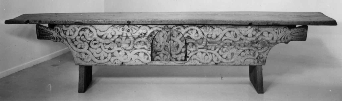 Langbord med forvaringsrom bak høye sarger. Fremsarg er med heldekkende flatskurd ranker og drageornamentikk samt en luke til forvaringsrommet. 