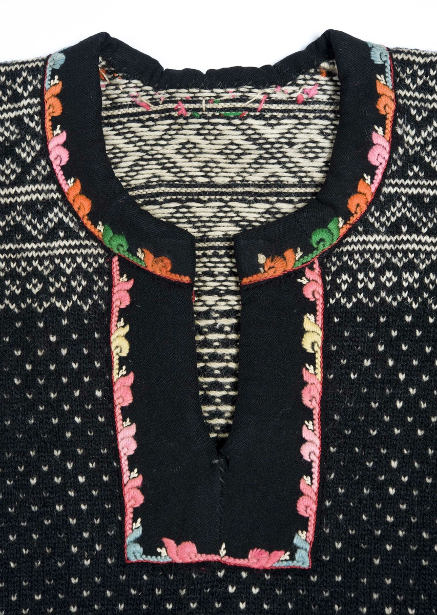 Strikket ullgarn, heimespunnet, svart og hvitt. Broderi i ullgarn. Langs nedrekanten en 13 cm bred hvit bord, resten mønsterstrikket: en bord åttebladroser, en sikksakkbord, en bord kors og kantstilte kvadrat. Langs åpningen foran og halsringingen er påsatt et stykke svart klede, saum i rosa, blått, gult og grønt zephyrgarn. Rundt ermet et 9,3 cm bredt belegg av svart klede, kantet med grønn klede 2,5 cm bred. En brodert bord langs indre og ytre kant av belegget. Aagot Noss 1969. 
