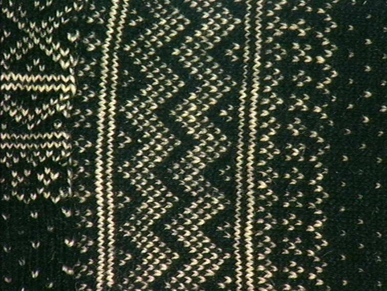 Strikket ullgarn, heimespunnet, svart og hvitt. Broderi i ullgarn. Langs nedrekanten en 13 cm bred hvit bord, resten mønsterstrikket: en bord åttebladroser, en sikksakkbord, en bord kors og kantstilte kvadrat. Langs åpningen foran og halsringingen er påsatt et stykke svart klede, saum i rosa, blått, gult og grønt zephyrgarn. Rundt ermet et 9,3 cm bredt belegg av svart klede, kantet med grønn klede 2,5 cm bred. En brodert bord langs indre og ytre kant av belegget. Aagot Noss 1969. 
